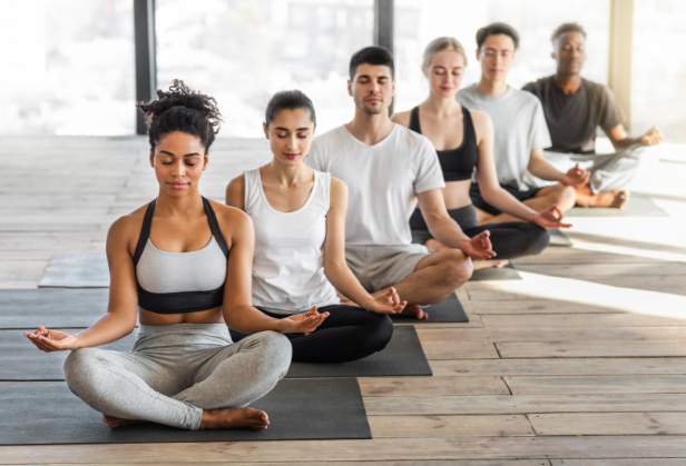 Six people practicing yoga in a bright studio, sitting cross-legged on mats with eyes closed, meditating.
