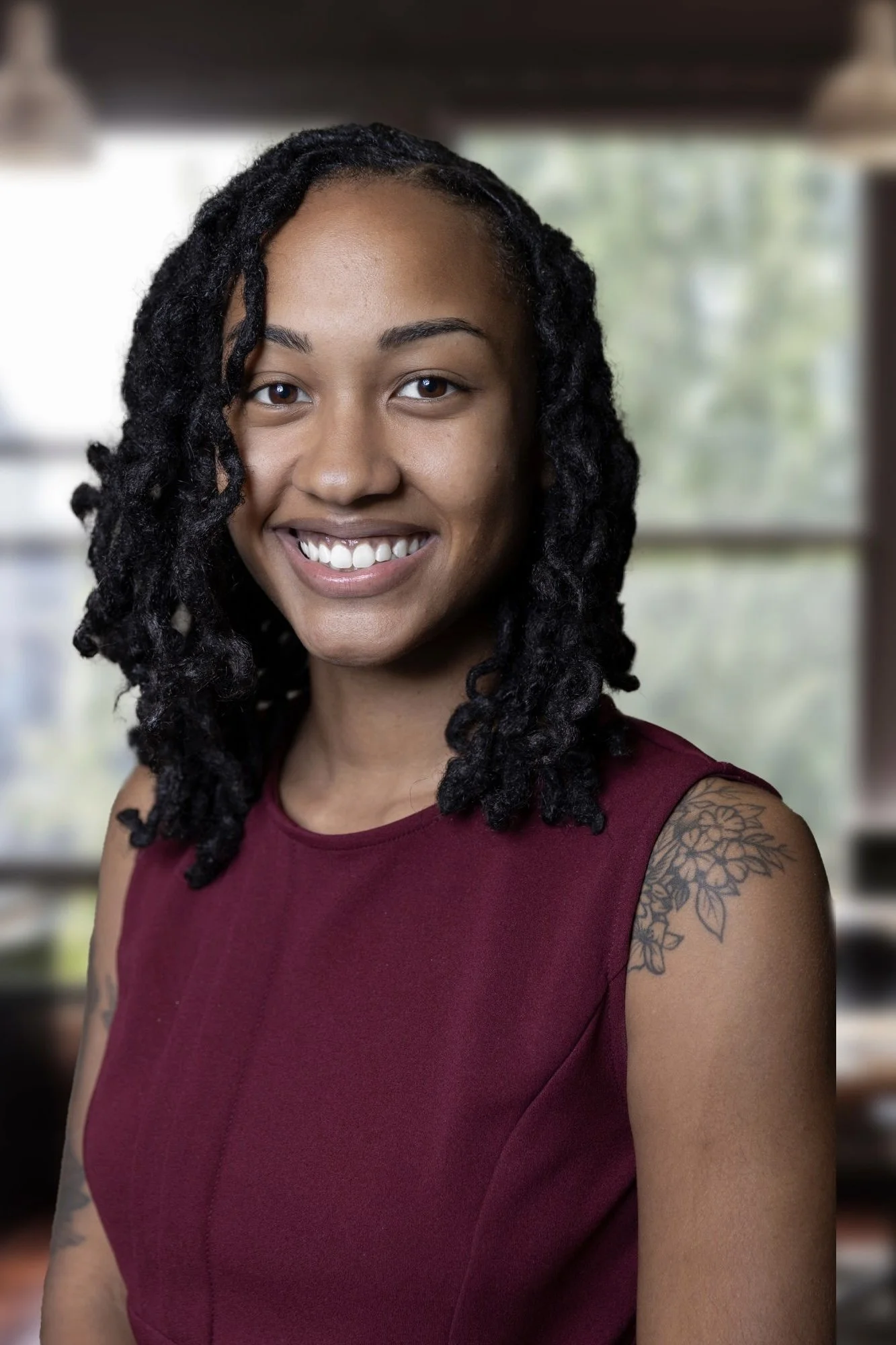 A young woman with shoulder-length curly black hair, smiling, wearing a sleeveless maroon top, and tattoos on her left shoulder. The background appears to be a room with large windows showing greenery outside.