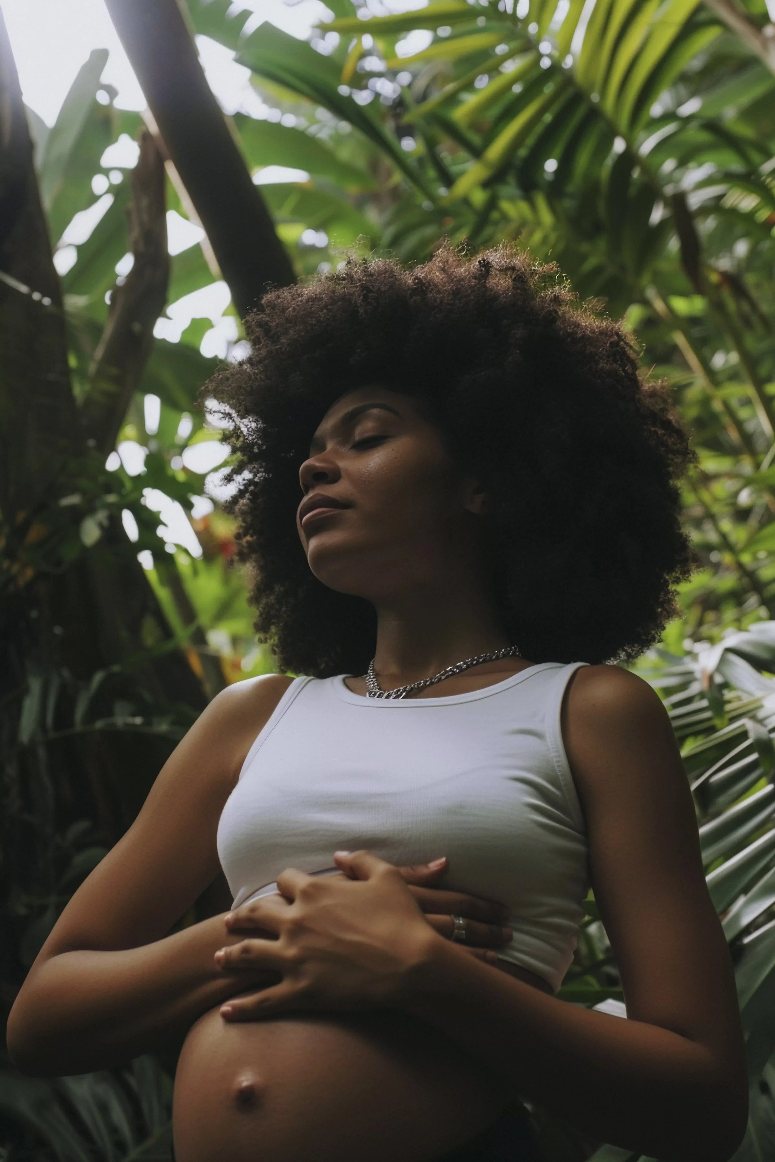 A pregnant woman with natural curly hair standing among lush green tropical plants with her eyes closed and arms crossed over her belly.