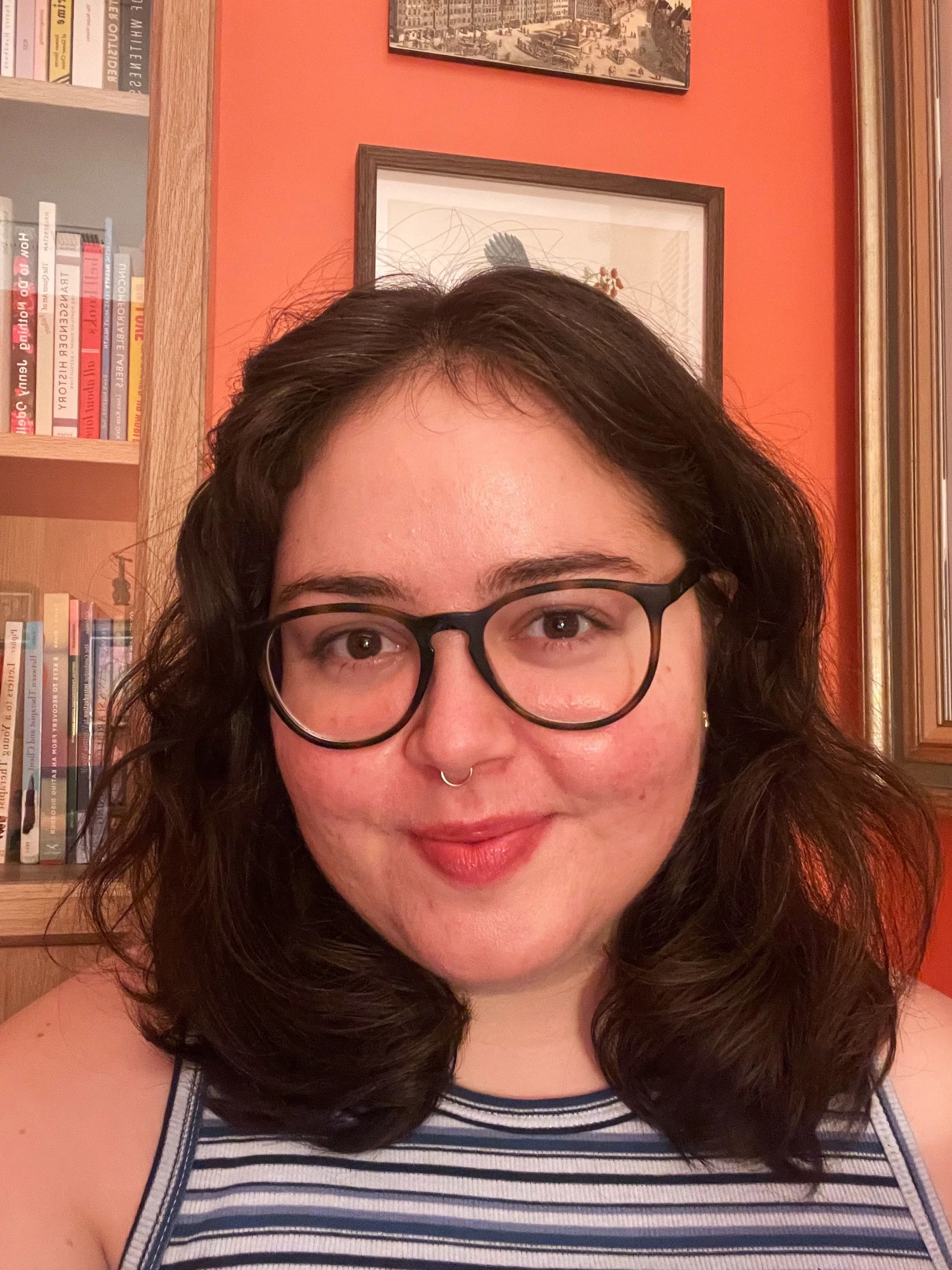 A nonbinary femme person with wavy brown hair, wearing glasses and a striped tank top, smiling in front of a bookshelf and framed art on a flamingo pink wall.