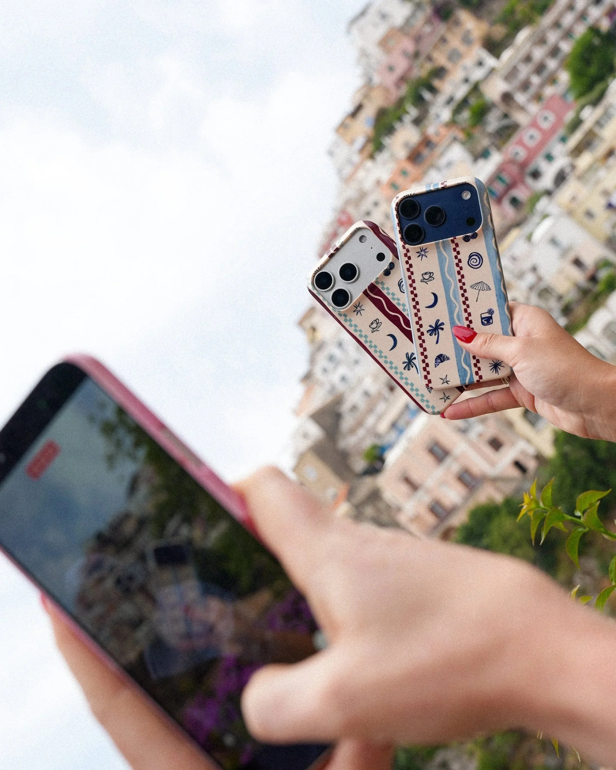A person with red nail polish holding two smartphones with colorful patterned cases, against a backdrop of apartment buildings and greenery on a cloudy day.