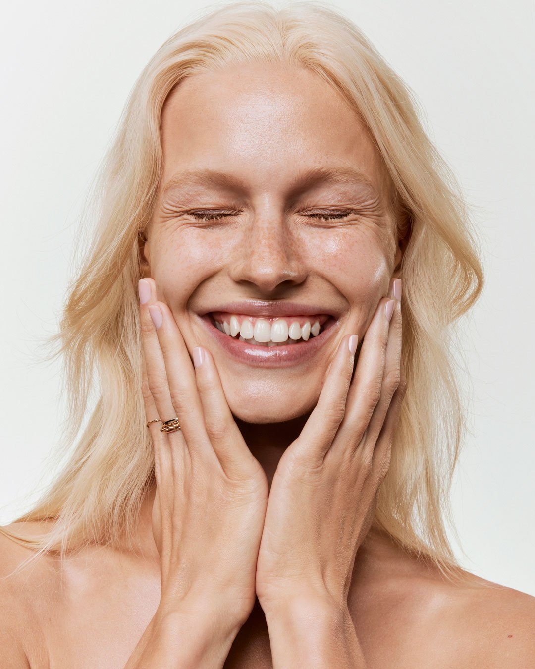 A woman with blonde hair smiling with her eyes closed, touching her cheeks with her hands.