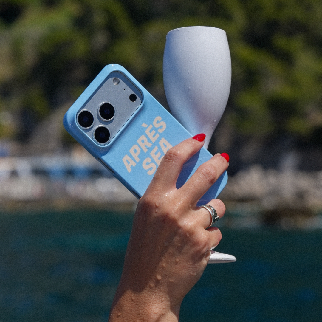 A person holding a blue phone with a case that says 'APRES SEA' and a white wine glass, with a background of water and greenery.