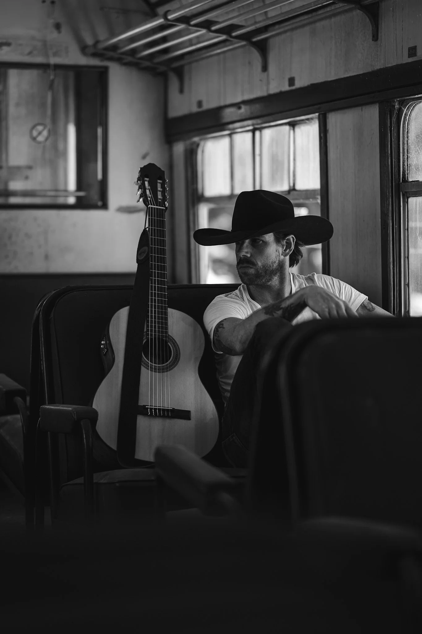 A man wearing a wide-brimmed hat sitting on a bus with a guitar resting against the seat next to him, looking out the window.
