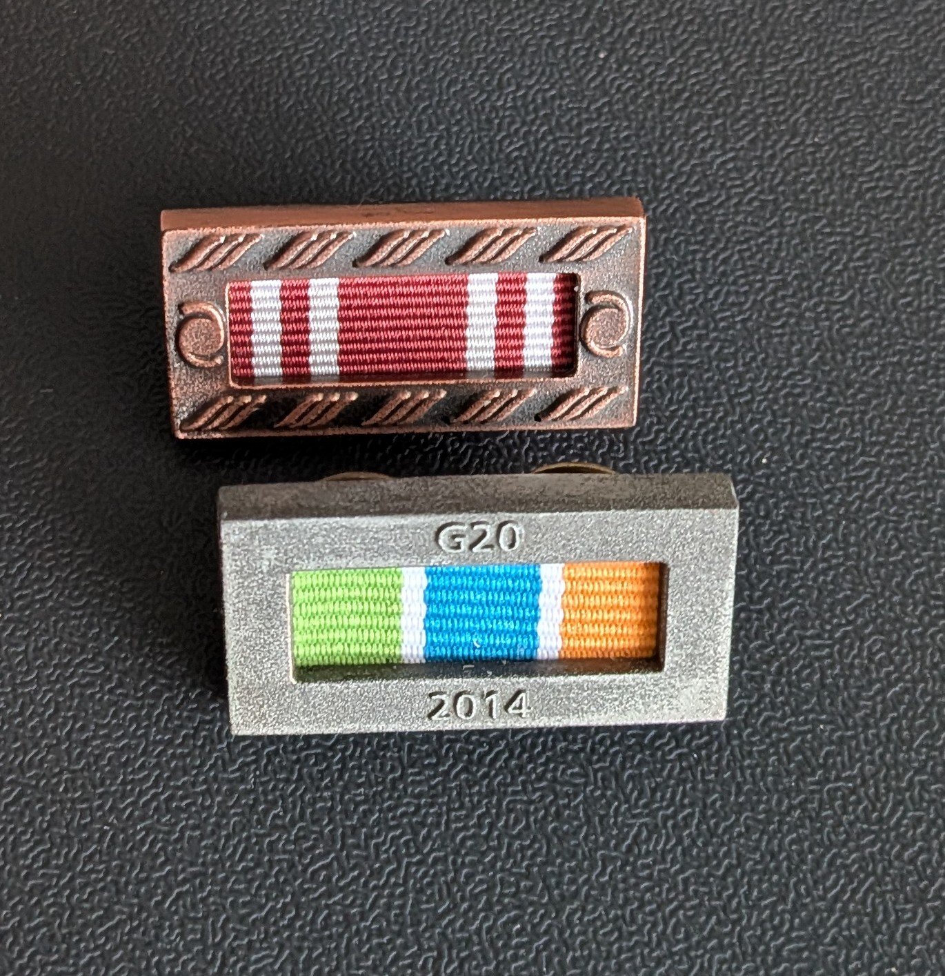 Two metal metal pins with colorful woven ribbons inside metal frames on a textured black surface.