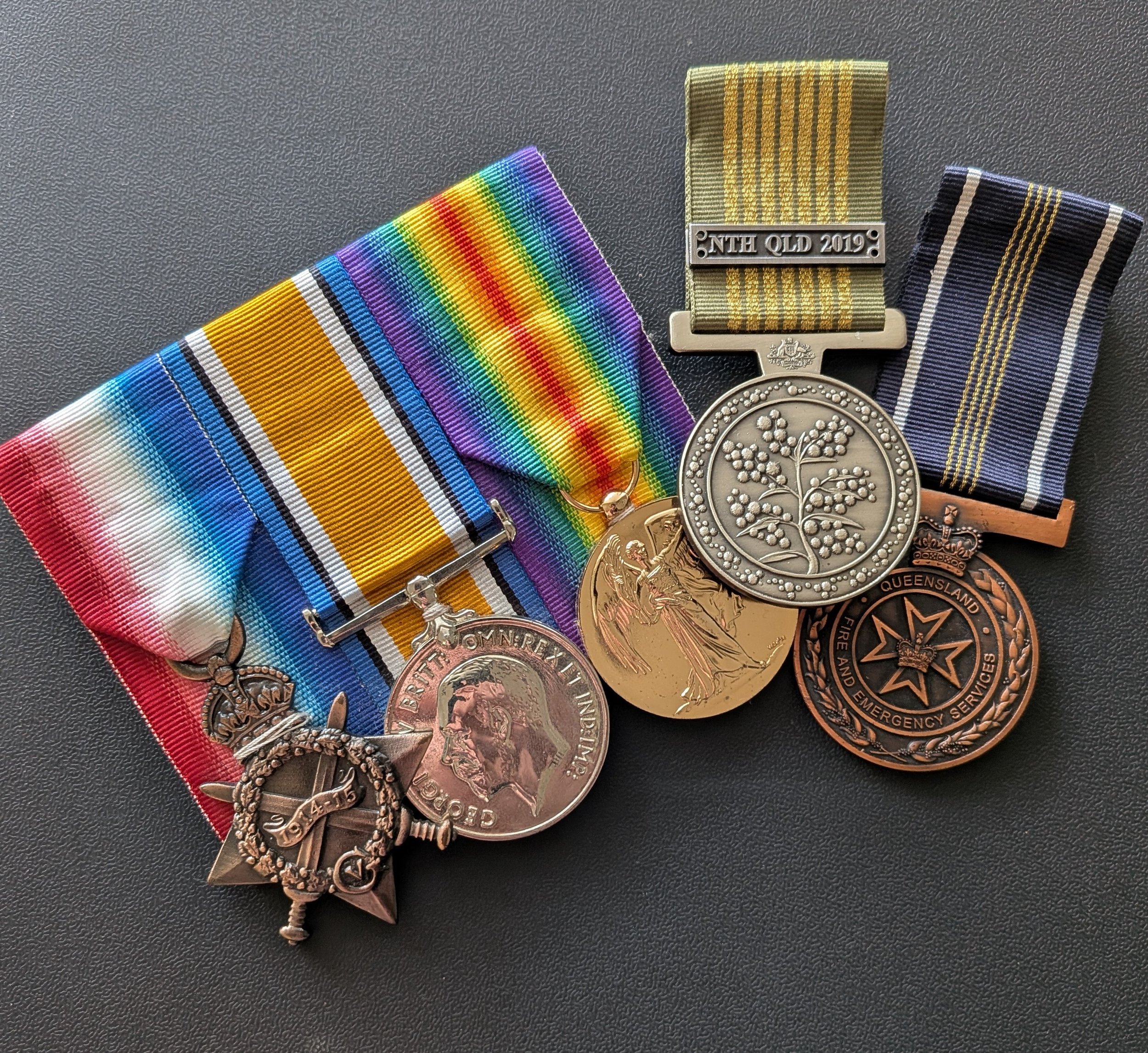 Close-up of several medals, including one with a rainbow ribbon, another with a military ribbon labeled "NTE QLD 2019," a third with a black and white striped ribbon, and a fourth with a badge that says "Queensland Fire and Emergency Services."