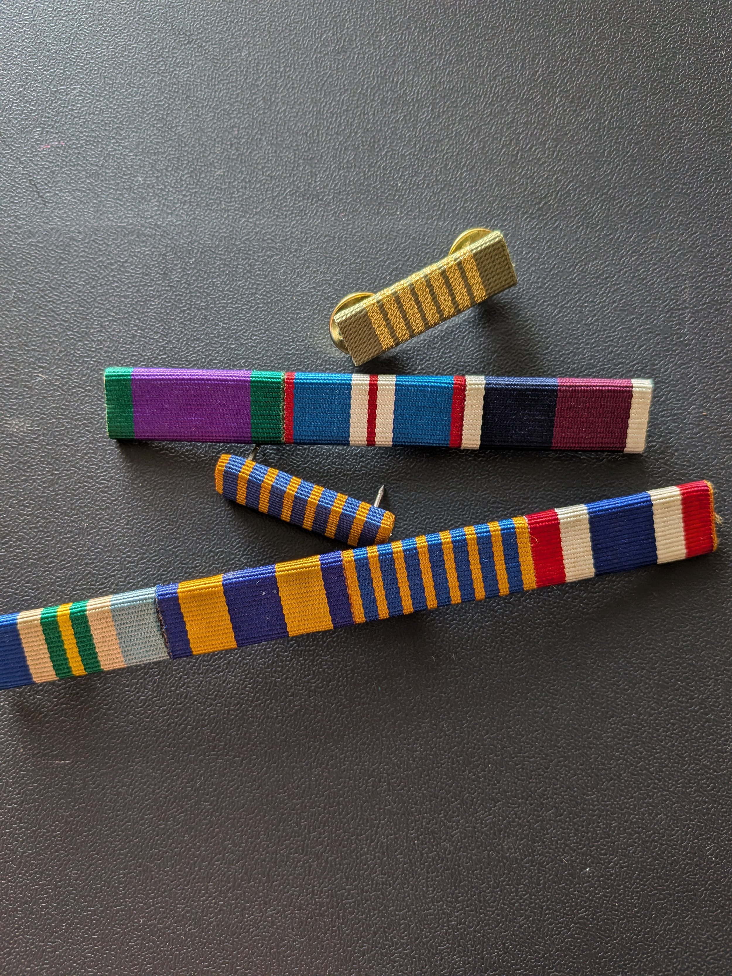 Close-up of several military medal ribbons and a gold-colored lapel pin on a dark textured surface.