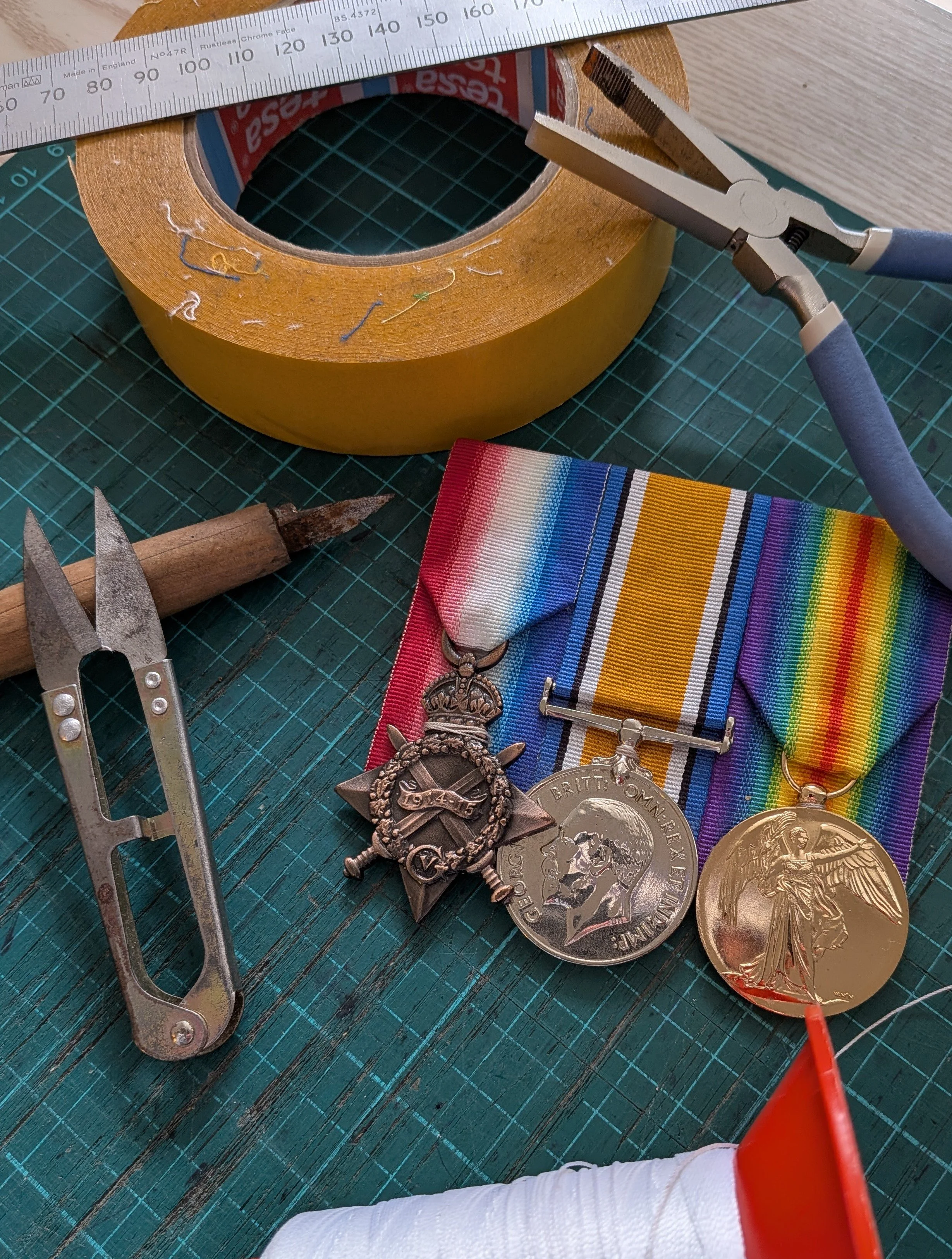 A roll of brown packing tape, a metal compass, a pair of scissors, military medals, and a rainbow-striped fabric on a green cutting mat.