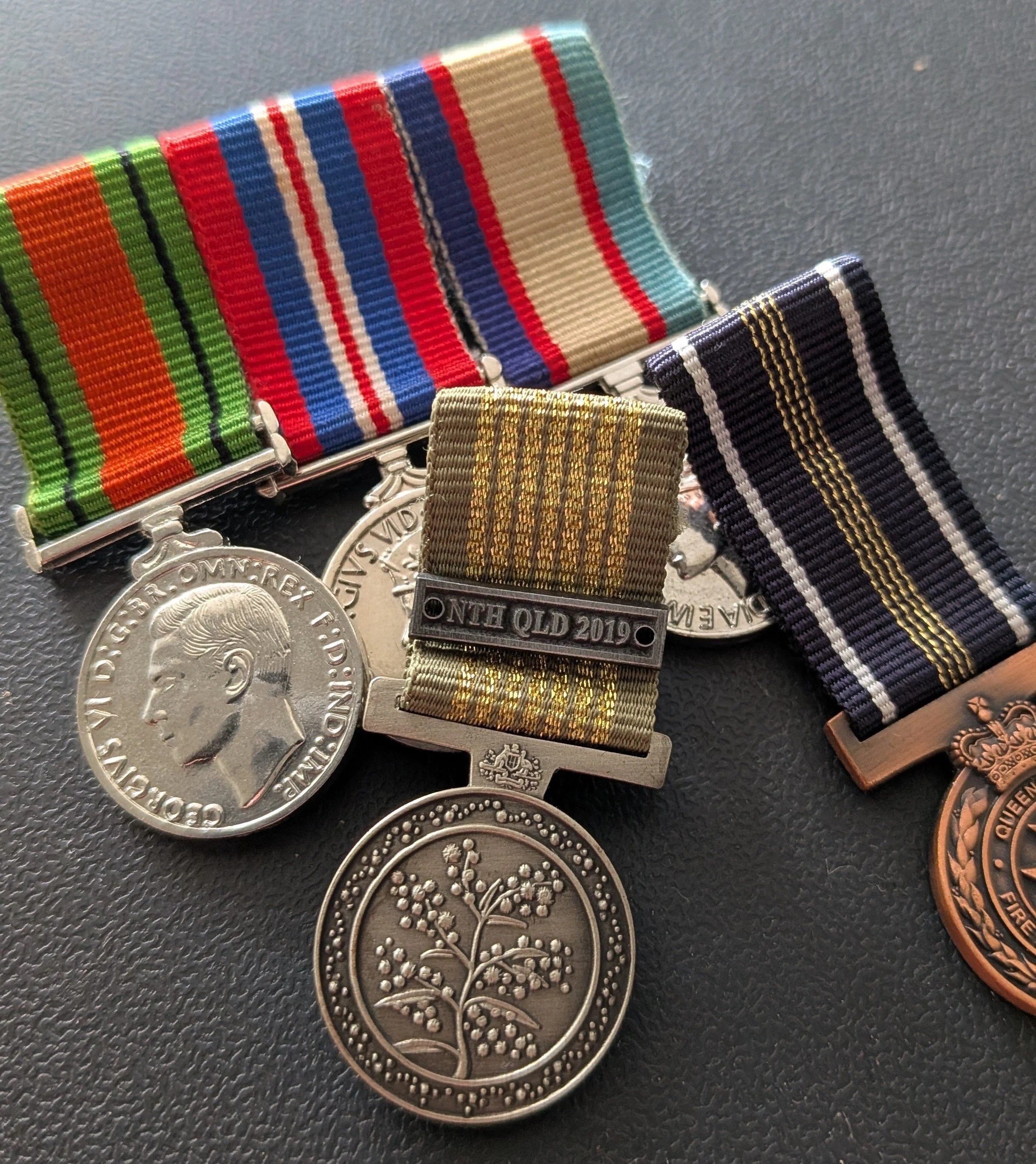 Various military medal ribbons and coins displayed on a black surface, including a silver coin with Queen Elizabeth II's profile and a coin with a tree design.