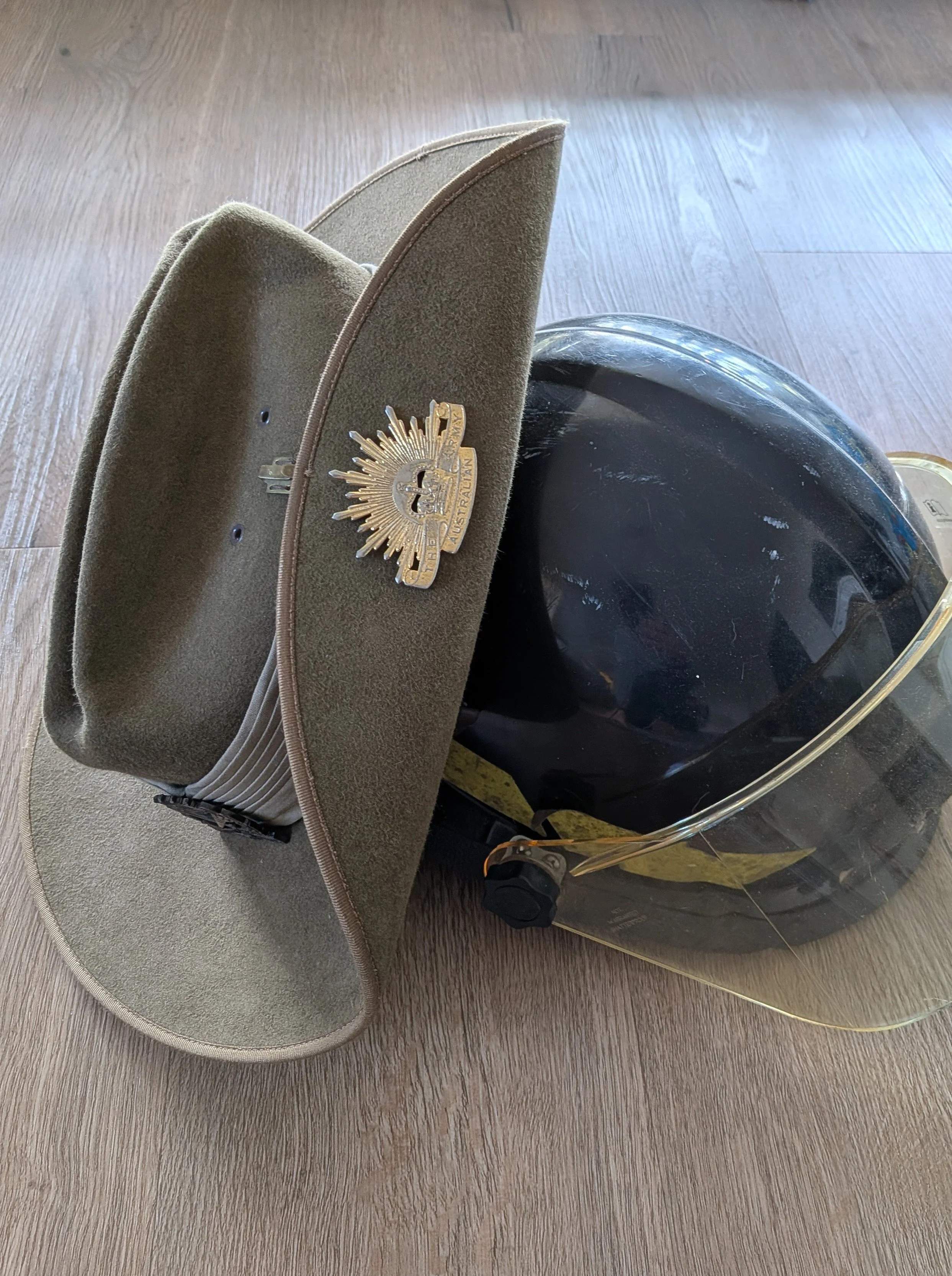 A military-style hat with a badge, a black motorcycle helmet, and a person’s foot in a black shoe on a wooden floor.