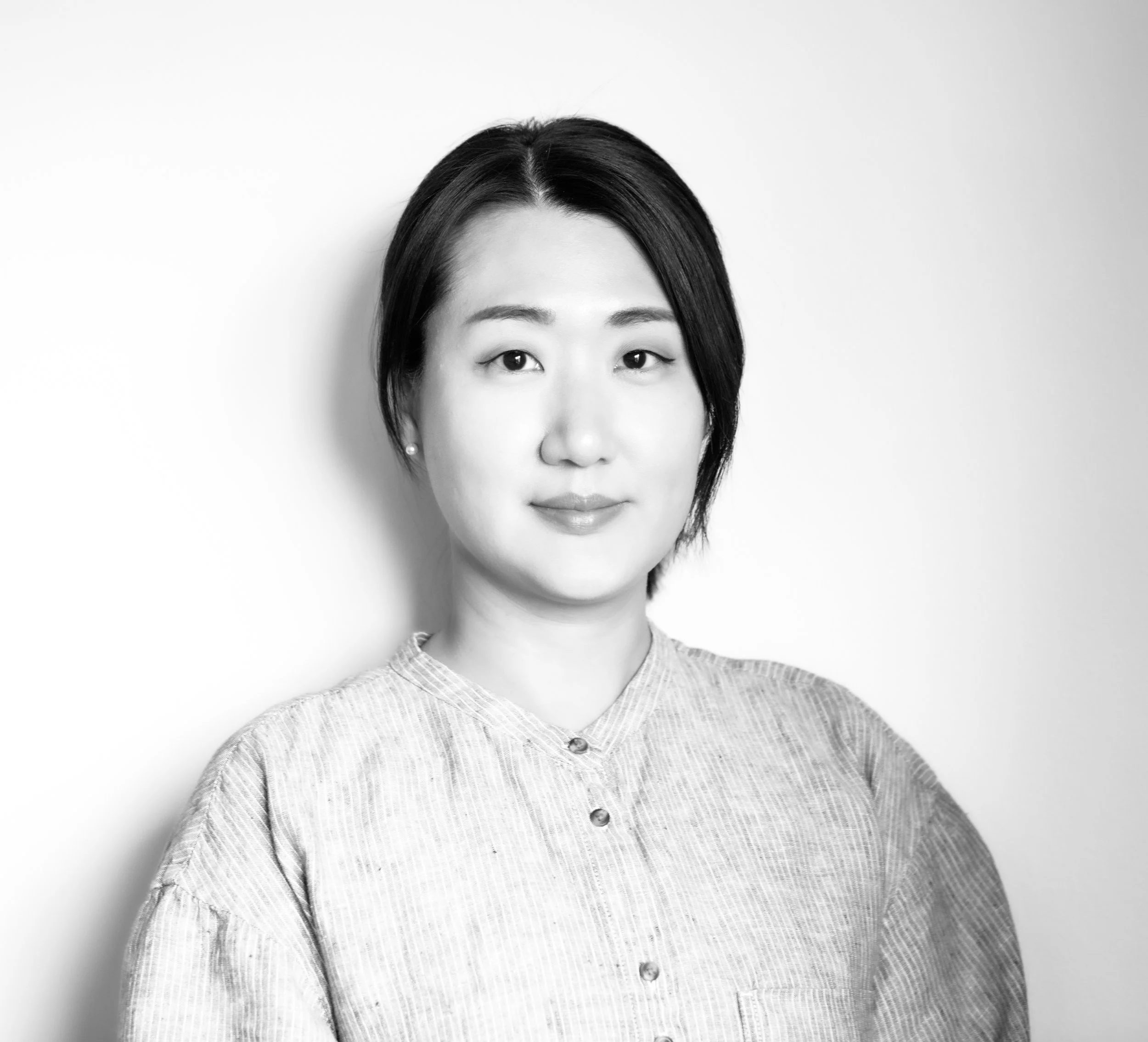 A black-and-white photo of a woman with short hair wearing a button-up shirt and pearl earrings, smiling softly and facing the camera against a plain white background.