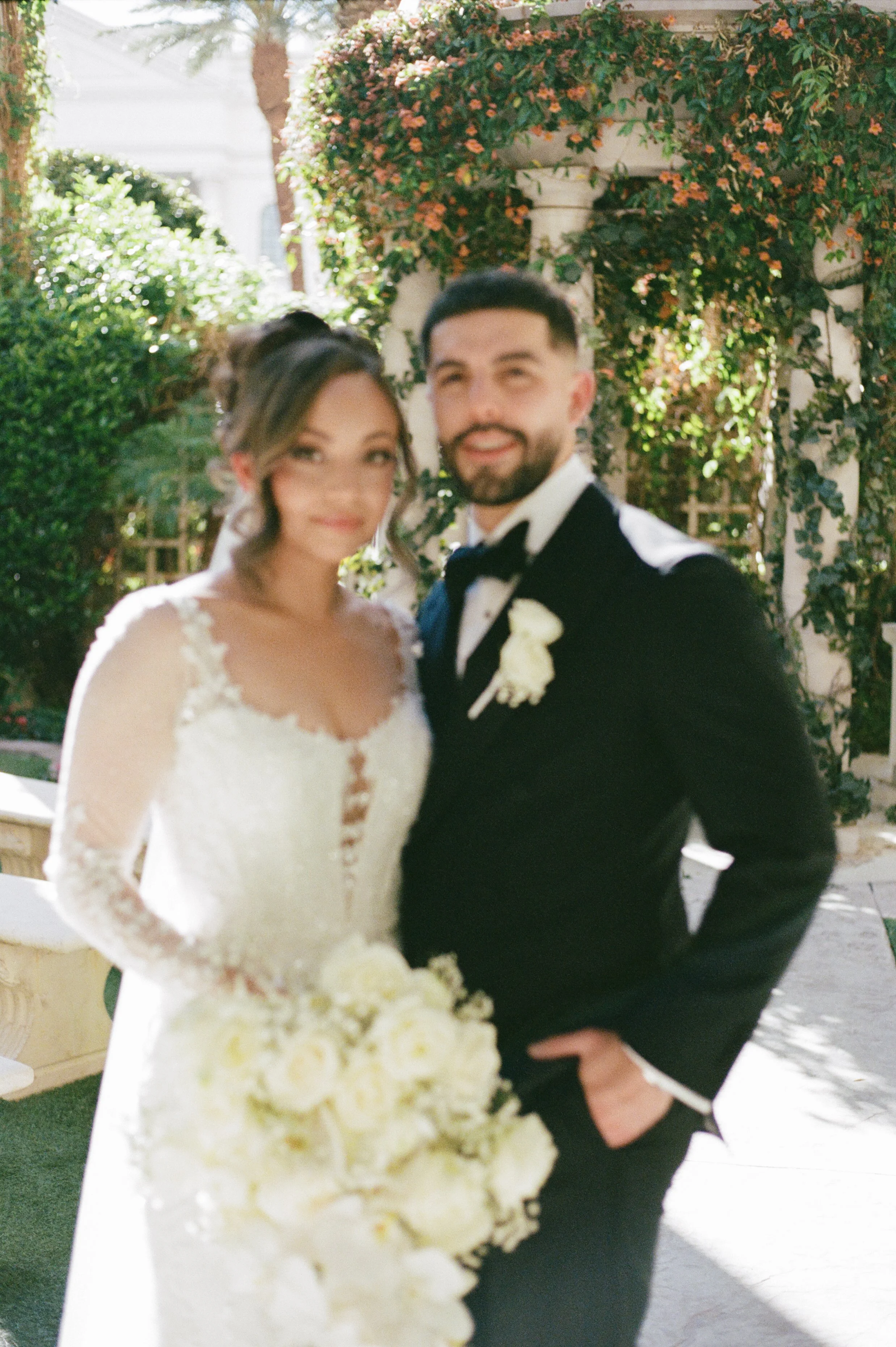 A bride and groom standing outdoors amid greenery, dressed in wedding attire, smiling and holding a bouquet of white flowers.