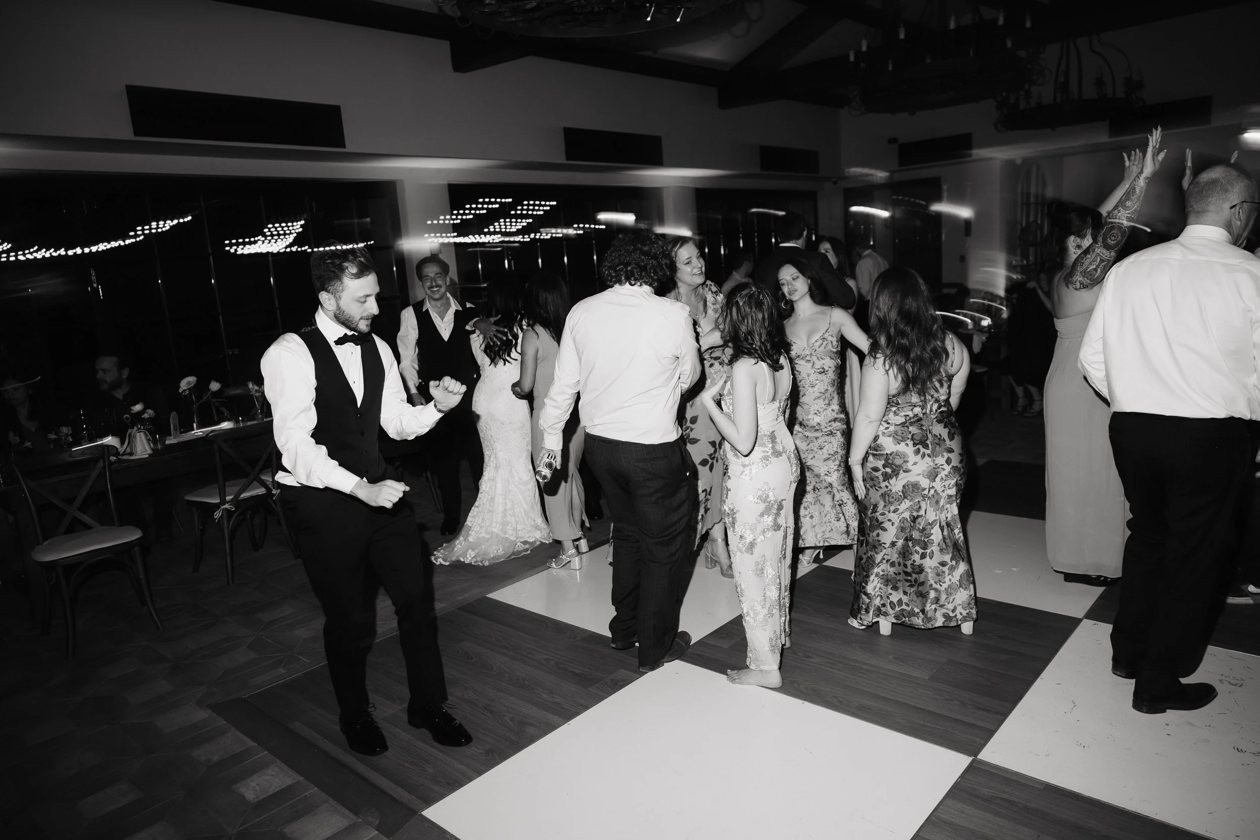 People dancing at a party in a modern indoor venue with a checkered dance floor, some wearing formal attire, black and white photo