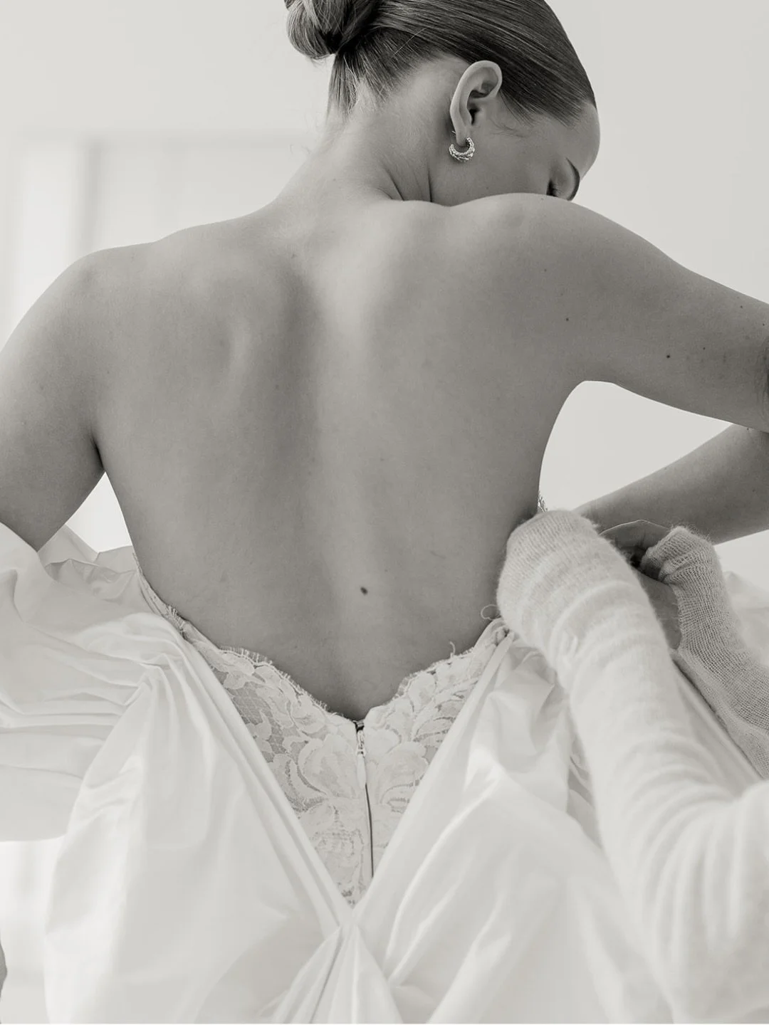 A woman with her back to the camera, being dressed in a wedding gown by another person wearing gloves.