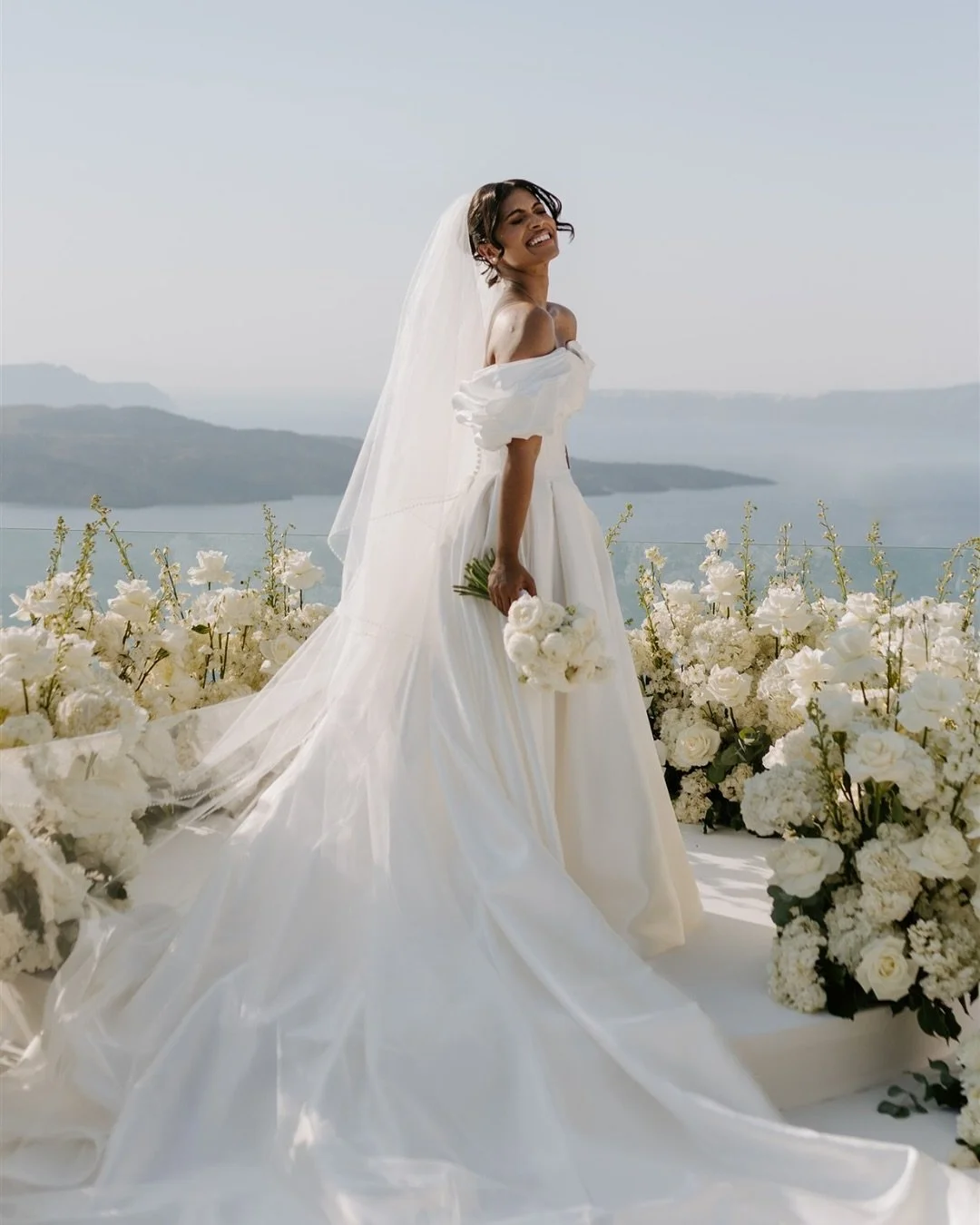 A bride in a white wedding dress holding a bouquet of white flowers, standing outdoors amidst white floral arrangements, with a scenic view of water and hills in the background.