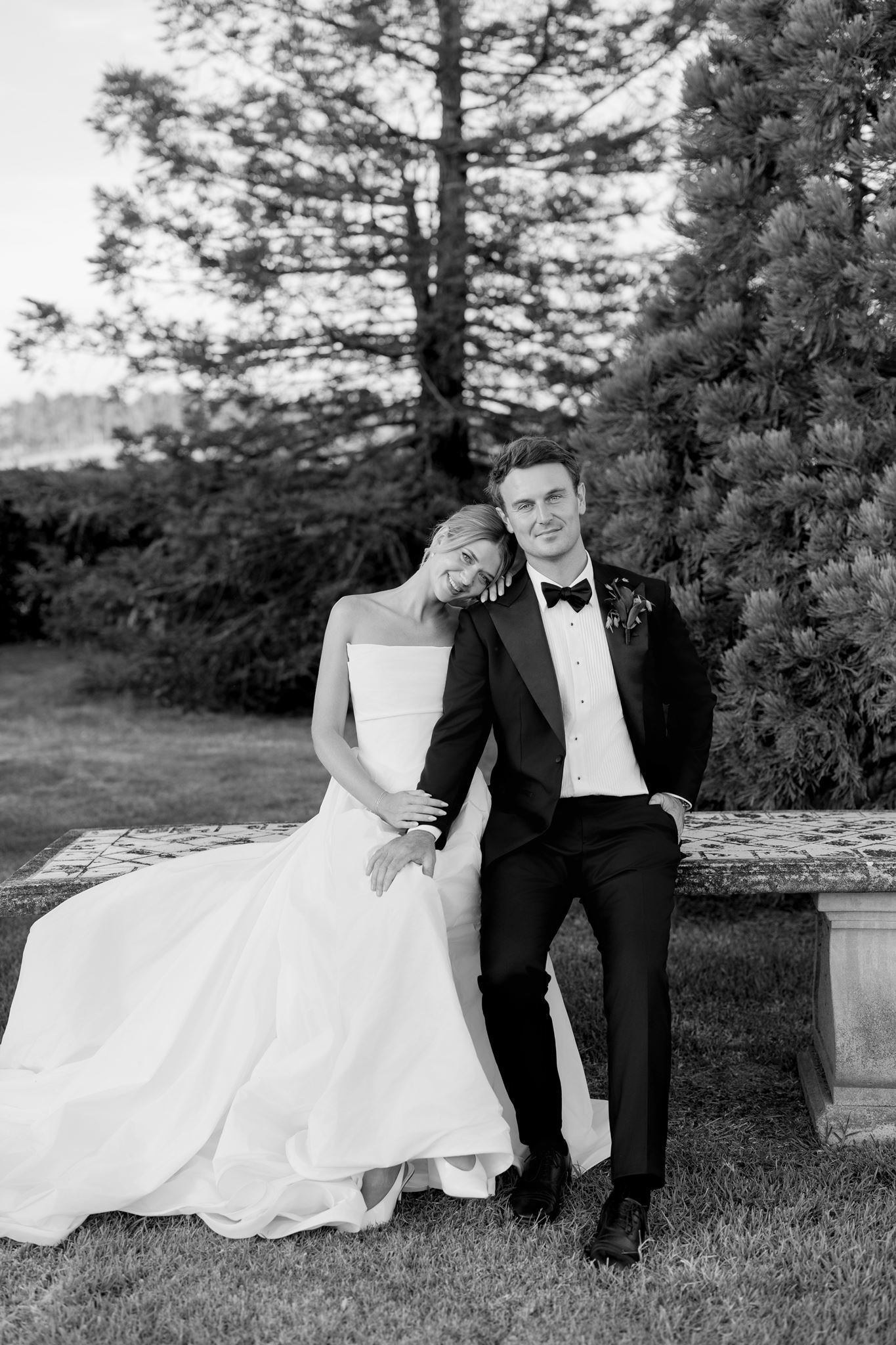 Black and white photo of a bride in a wedding gown and a groom in a tuxedo sitting on a bench outdoors. The bride is smiling with her head resting on the groom's shoulder, and the groom is looking at the camera. There are trees and bushes in the background.