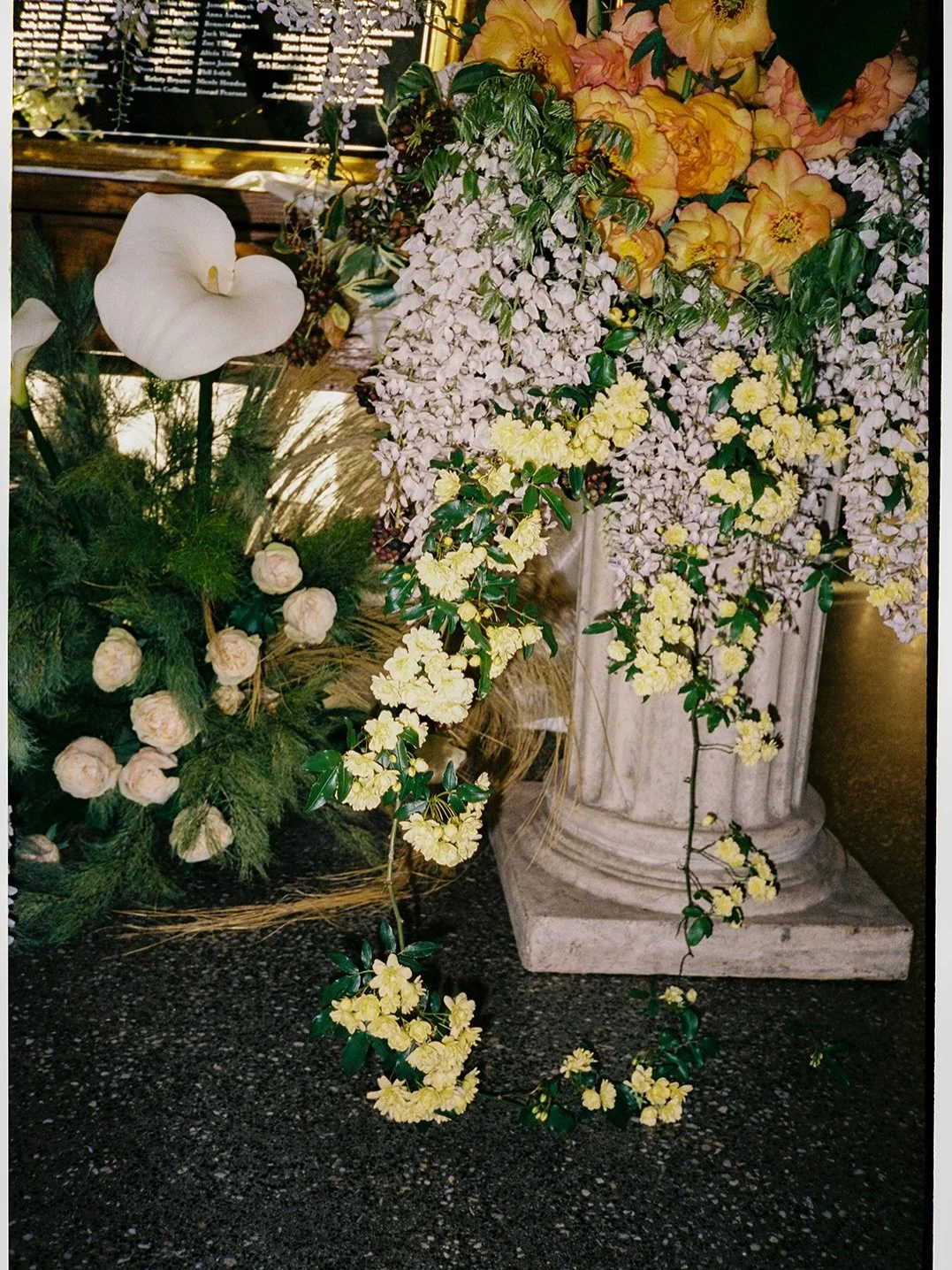 A floral arrangement with a white calla lily, cream roses, yellow flowers, and purple wisteria, displayed in a marble urn.