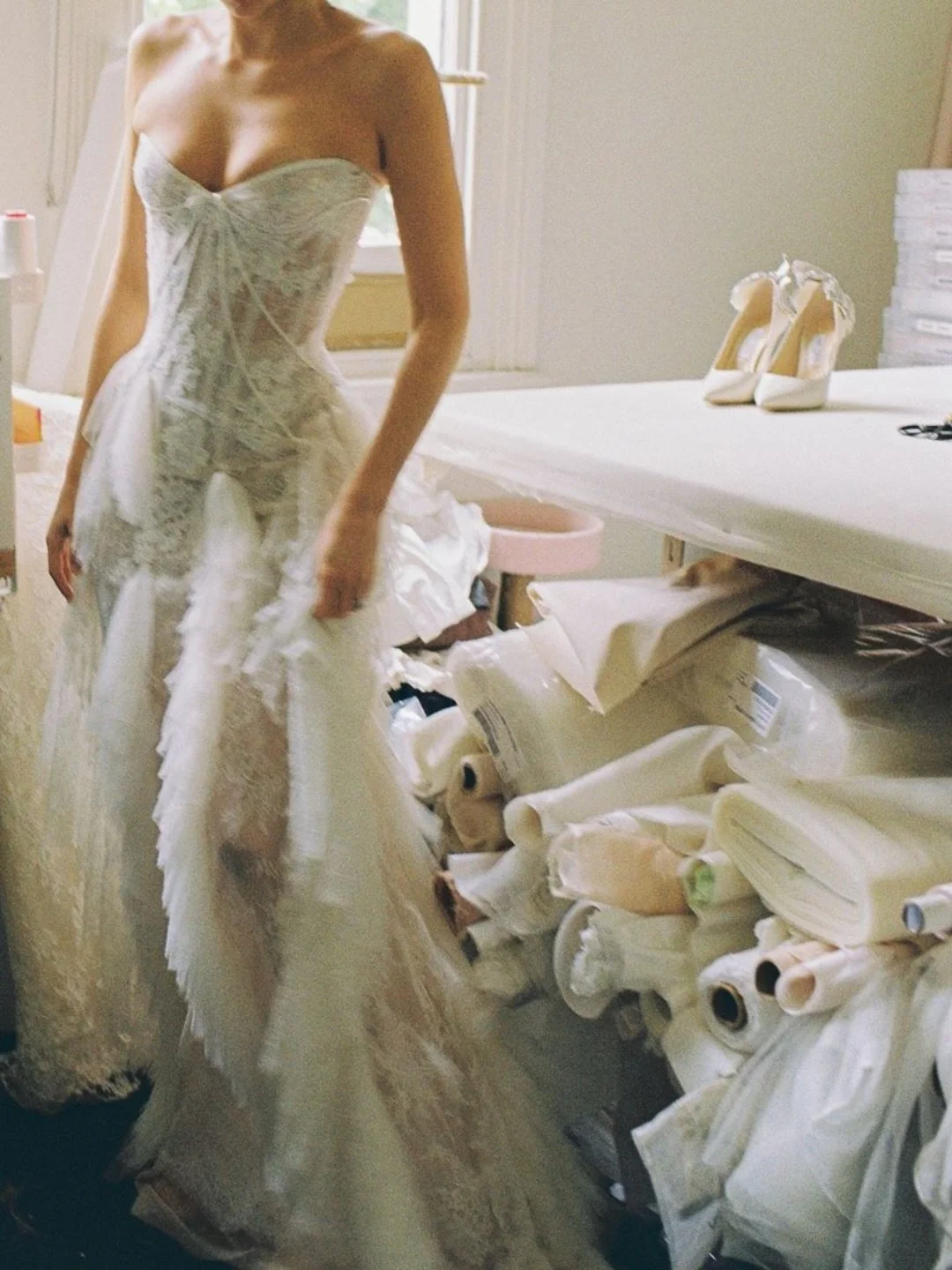 A woman in a white lace wedding dress standing in a room with a table holding white high-heeled shoes, and a large pile of fabric and sewing supplies below the table.
