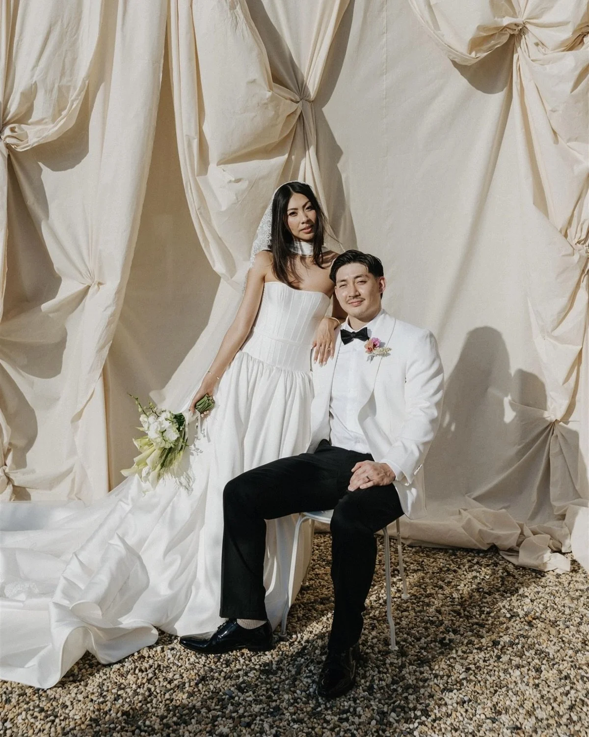 A couple dressed in wedding attire poses in front of a beige fabric backdrop. The bride is standing, wearing a strapless white wedding gown, holding a bouquet of white flowers, and has black hair. The groom is sitting on a chair, wearing a white tuxedo with black pants, a black bow tie, and has short black hair. They are both smiling slightly, and the bride's hand rests on the groom's shoulder.