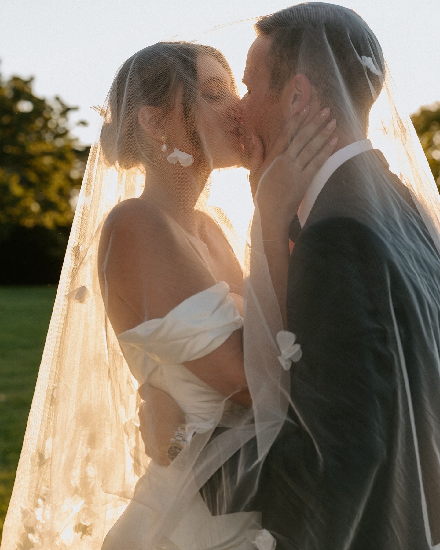 A bride and groom sharing a kiss outdoors during sunset. The bride is wearing a white dress and veil, and the groom is wearing a dark suit. They are holding each other's faces, with a backdrop of trees and a warm, glowing sky.