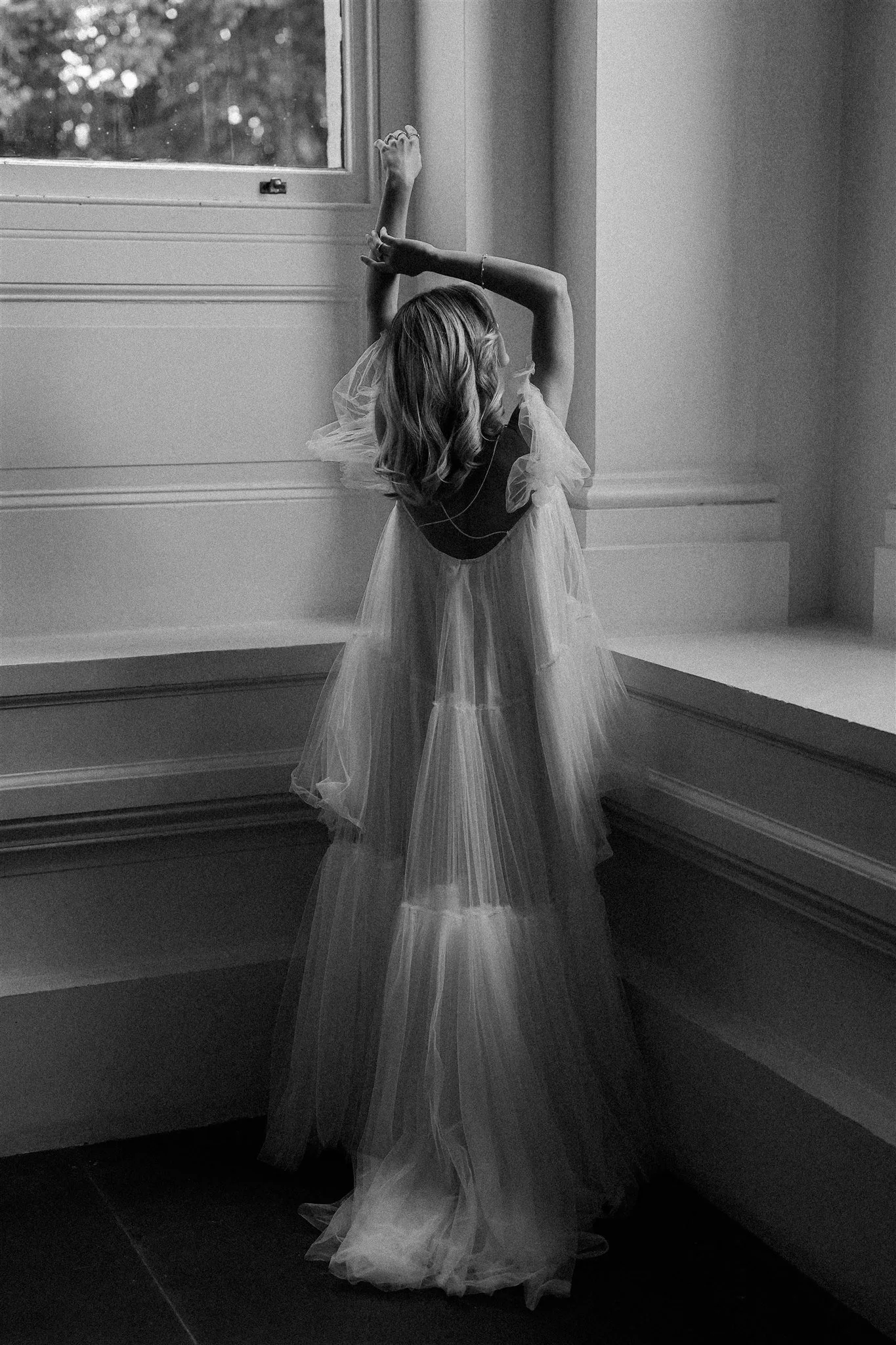 Black and white photo of a woman in a flowing gown standing by a large window, with her arms raised above her head and her face turned to the side.