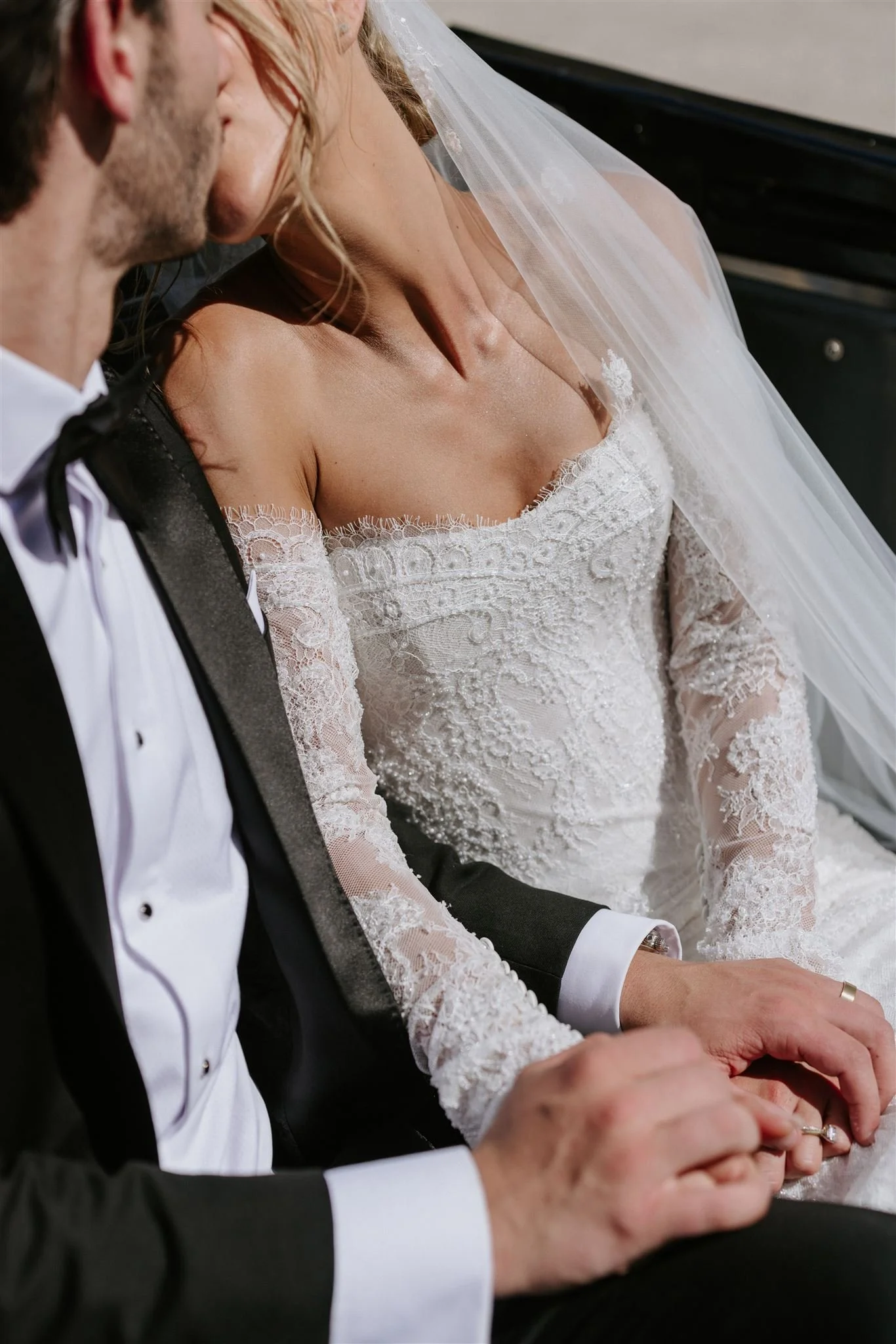 A close-up of a bride and groom holding hands, dressed in wedding attire, sitting close together.