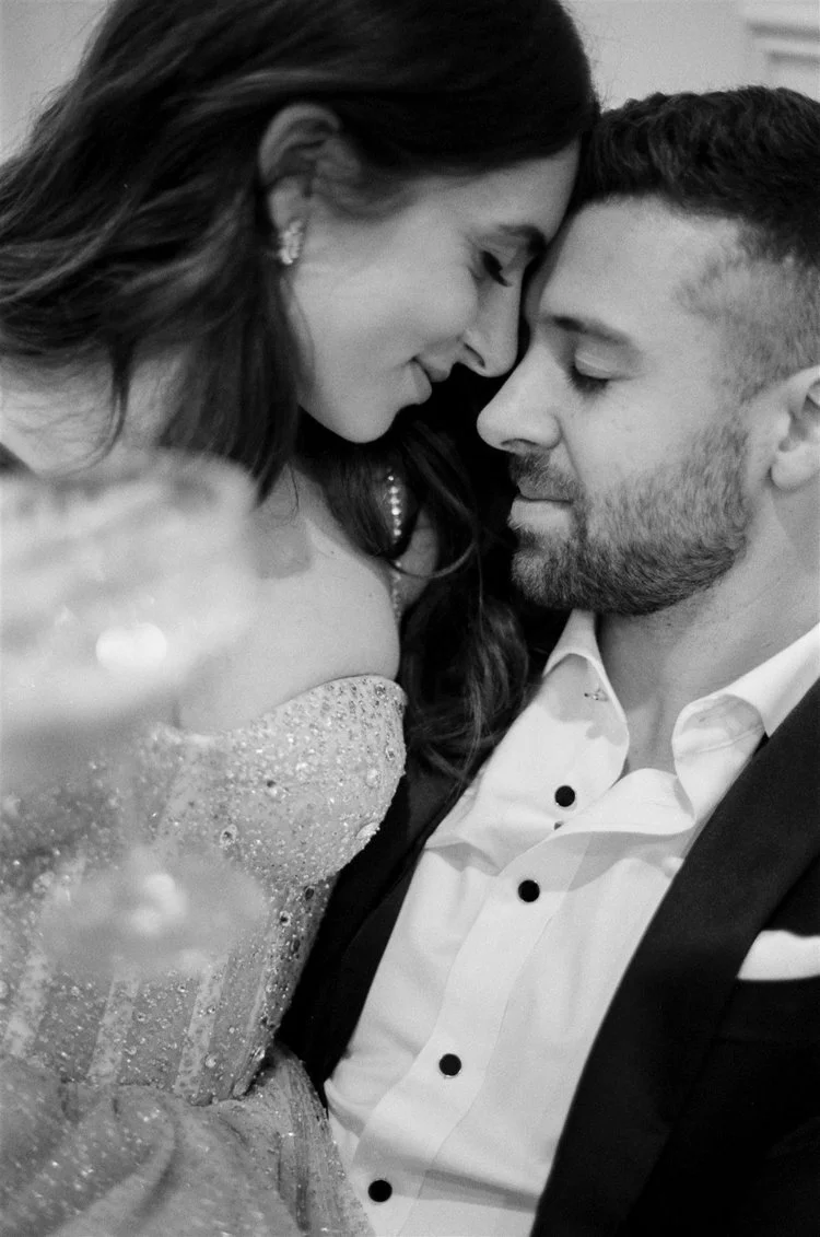 A black and white photo of a woman and man with their foreheads touching, both with closed eyes. The woman has long, dark hair and is wearing earrings and a sparkly dress, and the man has short hair and a beard, wearing a white shirt with black buttons and a dark jacket.