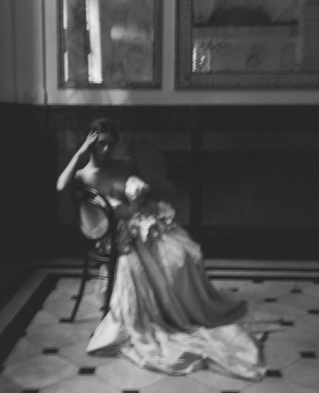 A black and white photograph of a woman sitting on a chair in a room with framed pictures on the wall. She is wearing a gown and has one hand touching her hair.