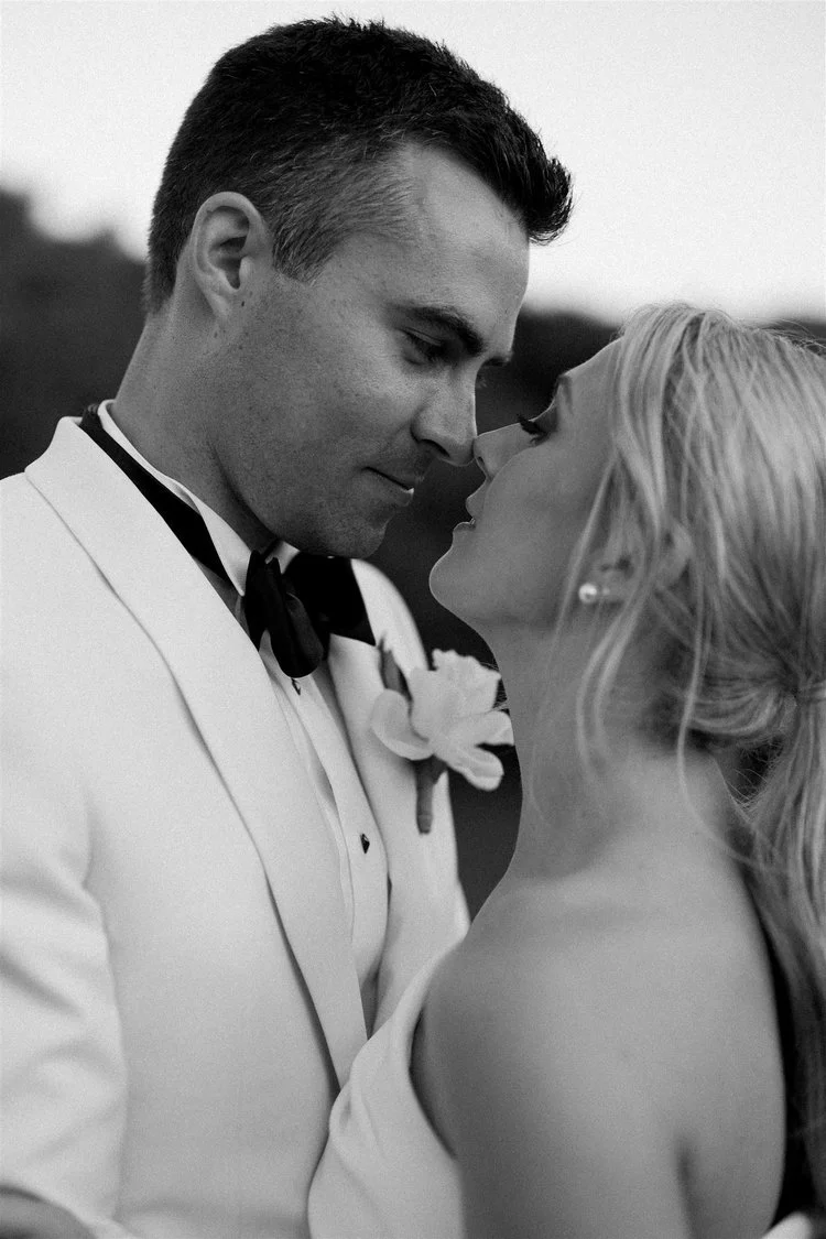 A black-and-white photo of a man and woman standing close, about to kiss, with their eyes closed. The man is wearing a tuxedo with a bow tie and a boutonniere, and the woman has blonde hair styled in loose curls and is wearing earrings.