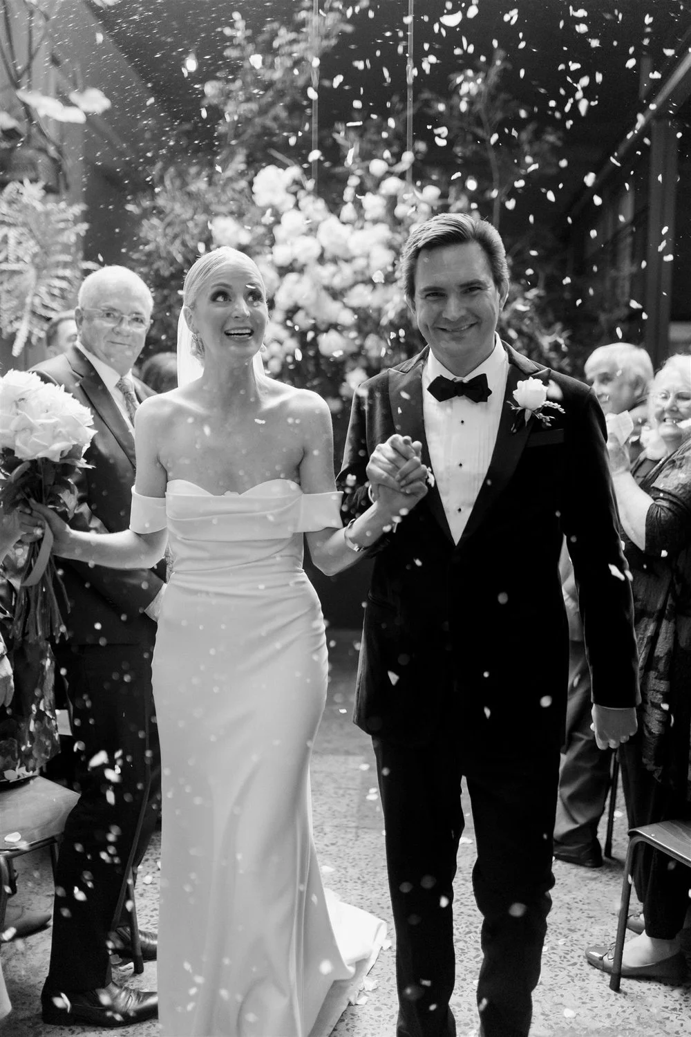 A black-and-white photo of a bride and groom holding hands and smiling at their wedding celebration, with guests and floral decorations in the background, as confetti falls around them.