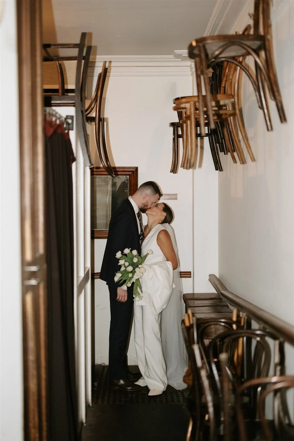 A bride and groom sharing a kiss in a narrow hallway, with stacked chairs hanging on the ceiling and wooden chairs along the walls, during their wedding.