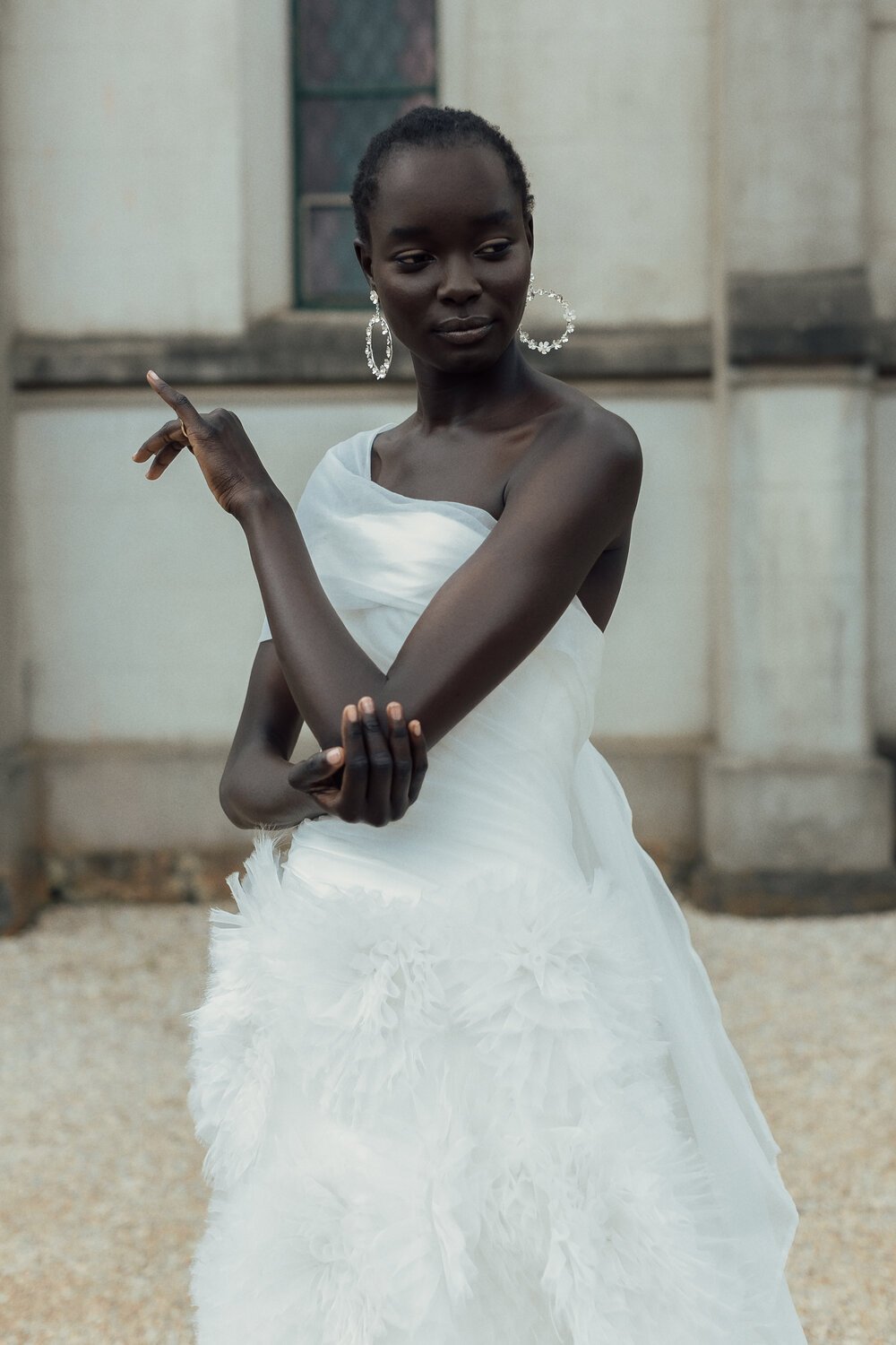 A woman in a white dress with feather details, wearing large hoop earrings, is posing outdoors in front of a building with beige walls and a window, with a confident expression and a gesture with her arm.