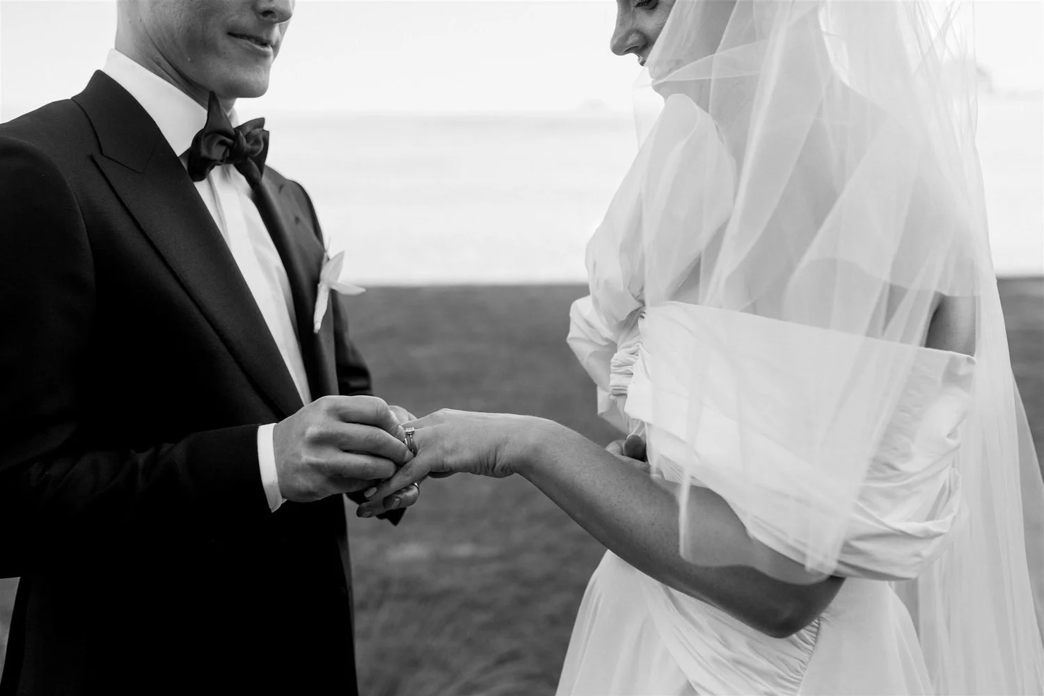 A black-and-white photo of a wedding ceremony showing a groom in a suit and tie holding a bride's hand as he places a ring on her finger, with the bride wearing a wedding dress and veil.