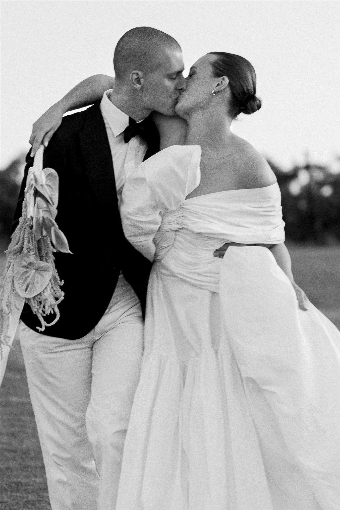 A black and white photo of a couple kissing. The man is dressed in formalwear, wearing a tuxedo with a bow tie. The woman is in a strapless wedding gown with draped fabric. She has her hair styled in an updo. The scene appears to be at a wedding or special event.