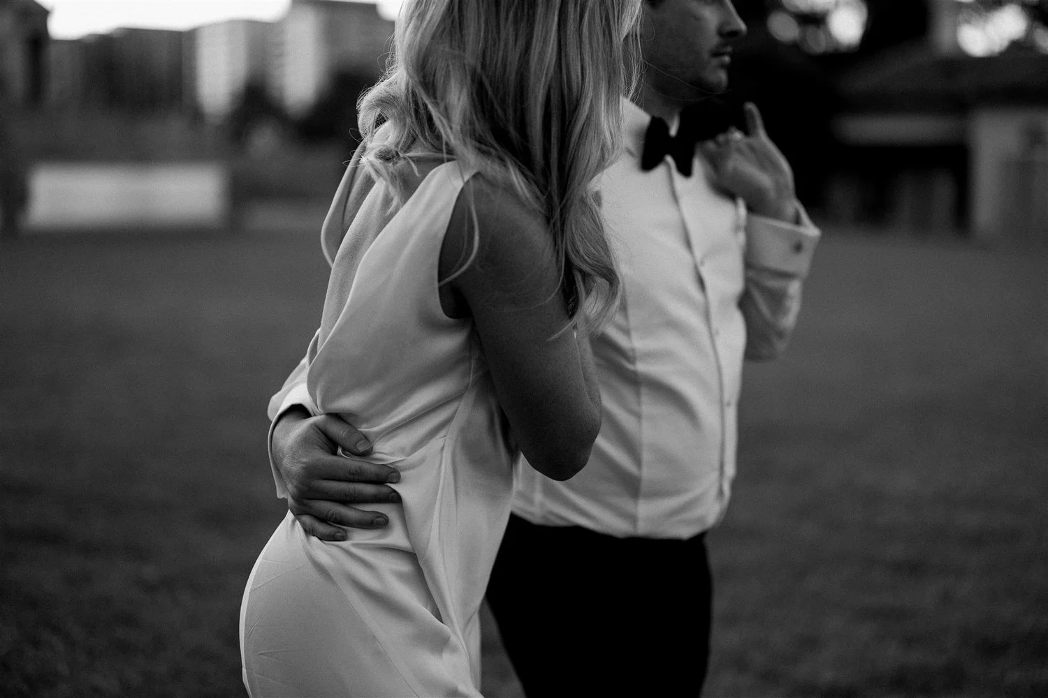 A black and white photo of a woman and a man standing close together outdoors, with the woman's hand on her hip and her arm around the man's shoulder, both dressed formally.