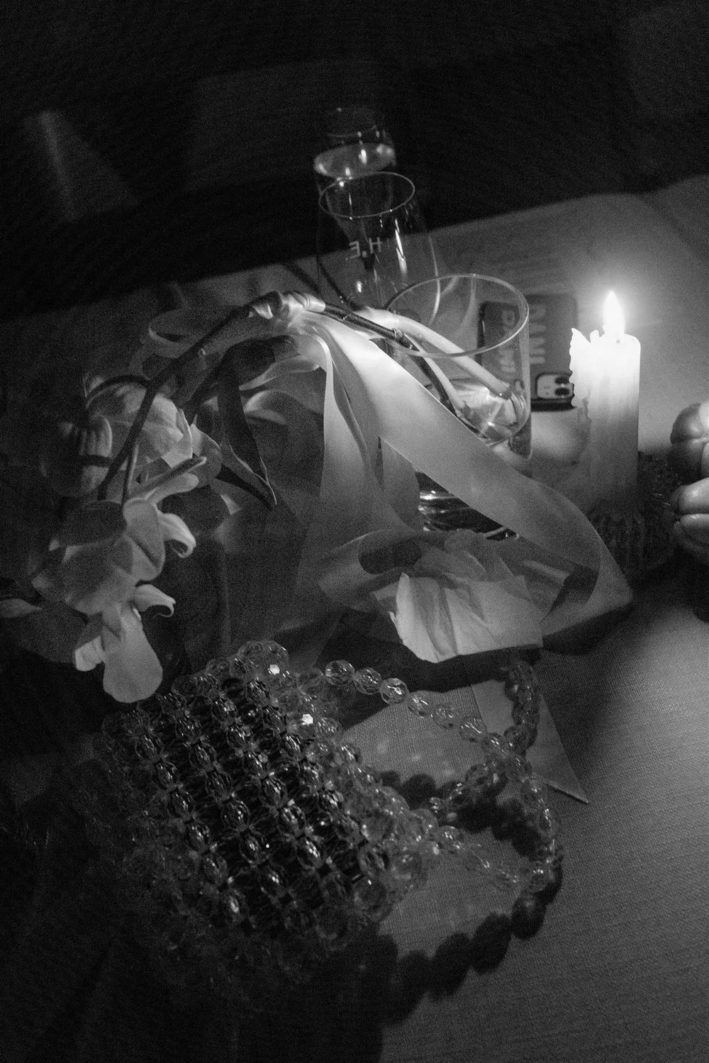 A table with flowers, glassware, a candle, a jewelry box, and a hand holding a pair of scissors in a dimly lit setting.
