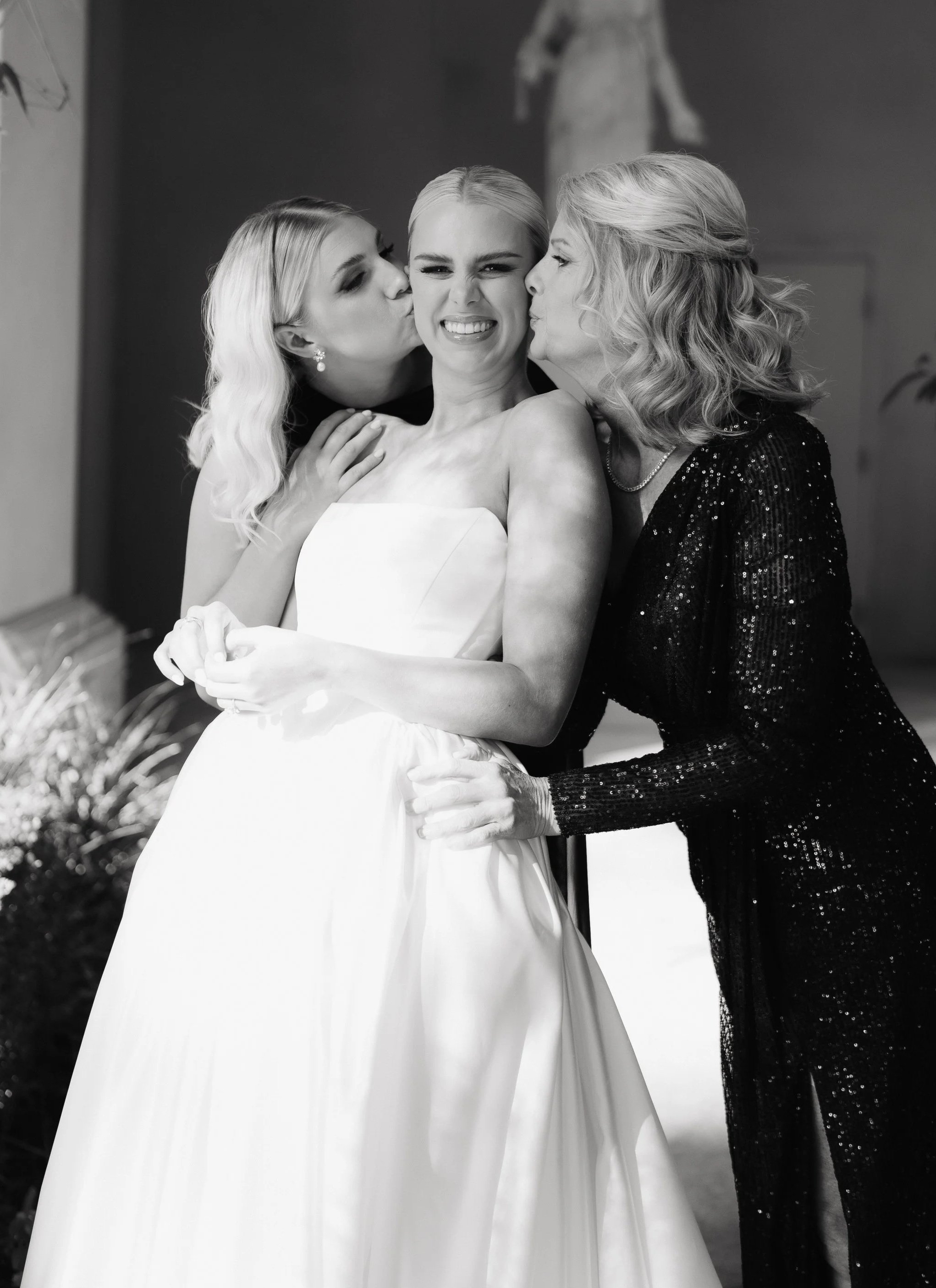 Three women, one in a wedding dress and two in formal attire, are kissing the bride on both cheeks while she smiles.