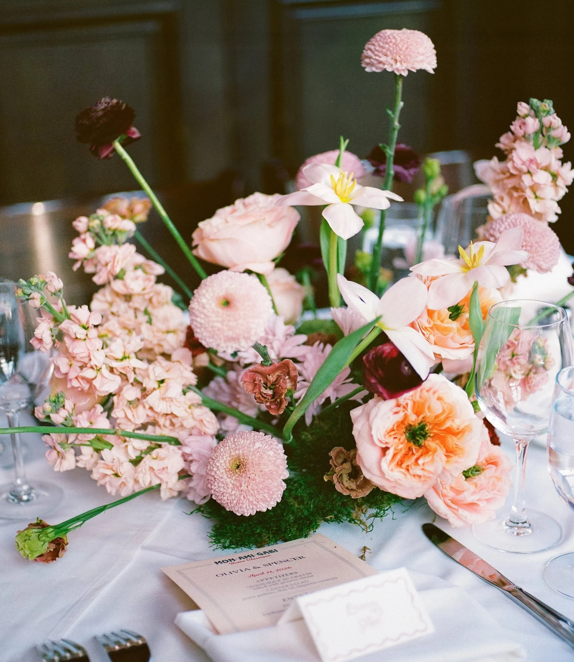 A floral centerpiece with pink and burgundy flowers on a white table with a menu card and silverware.