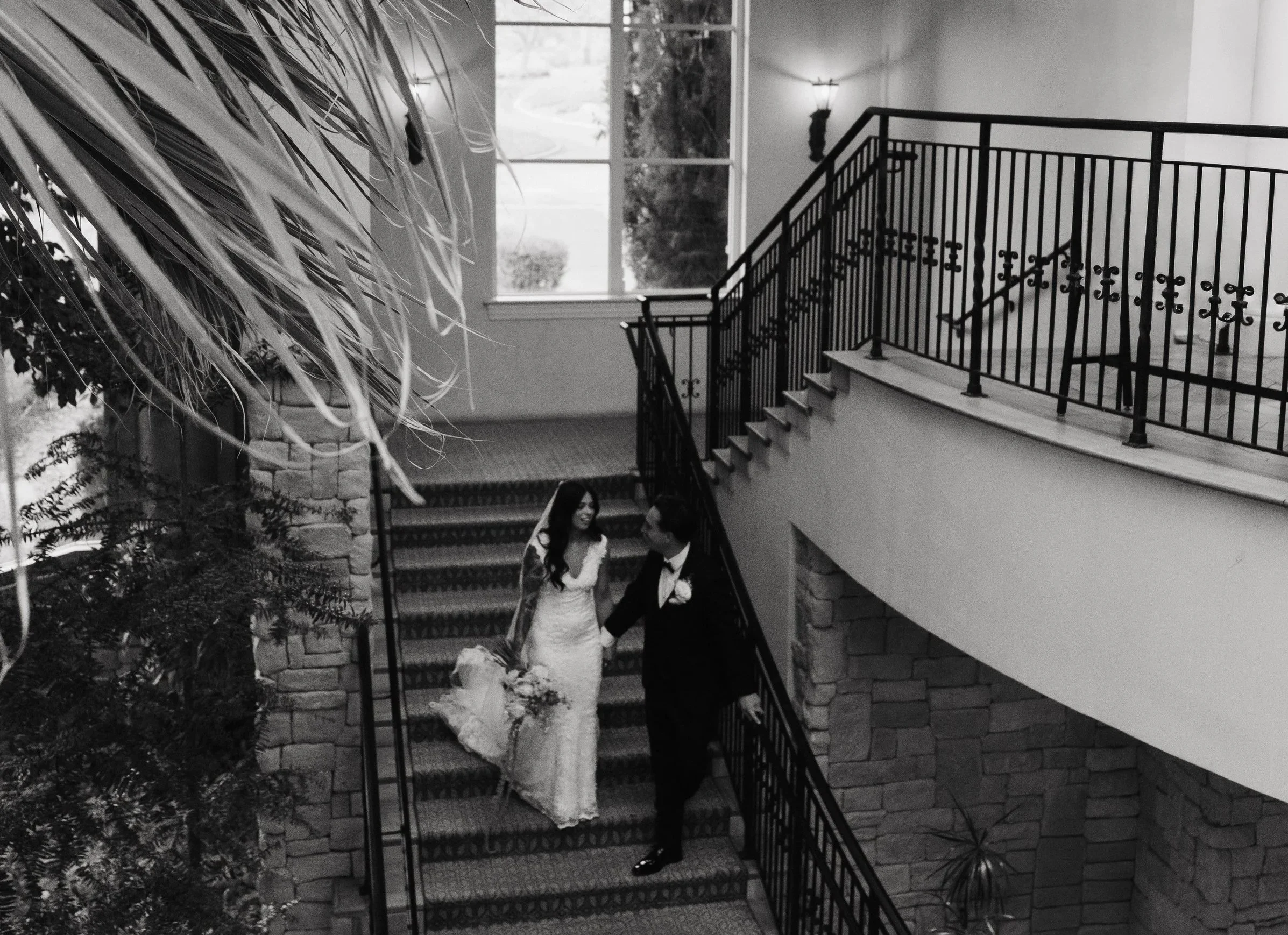 A bride and groom are standing on a staircase in a well-lit interior space, possibly during their wedding. The bride wears a long white wedding dress and is holding a bouquet, while the groom wears a dark tuxedo. They are smiling at each other, posit