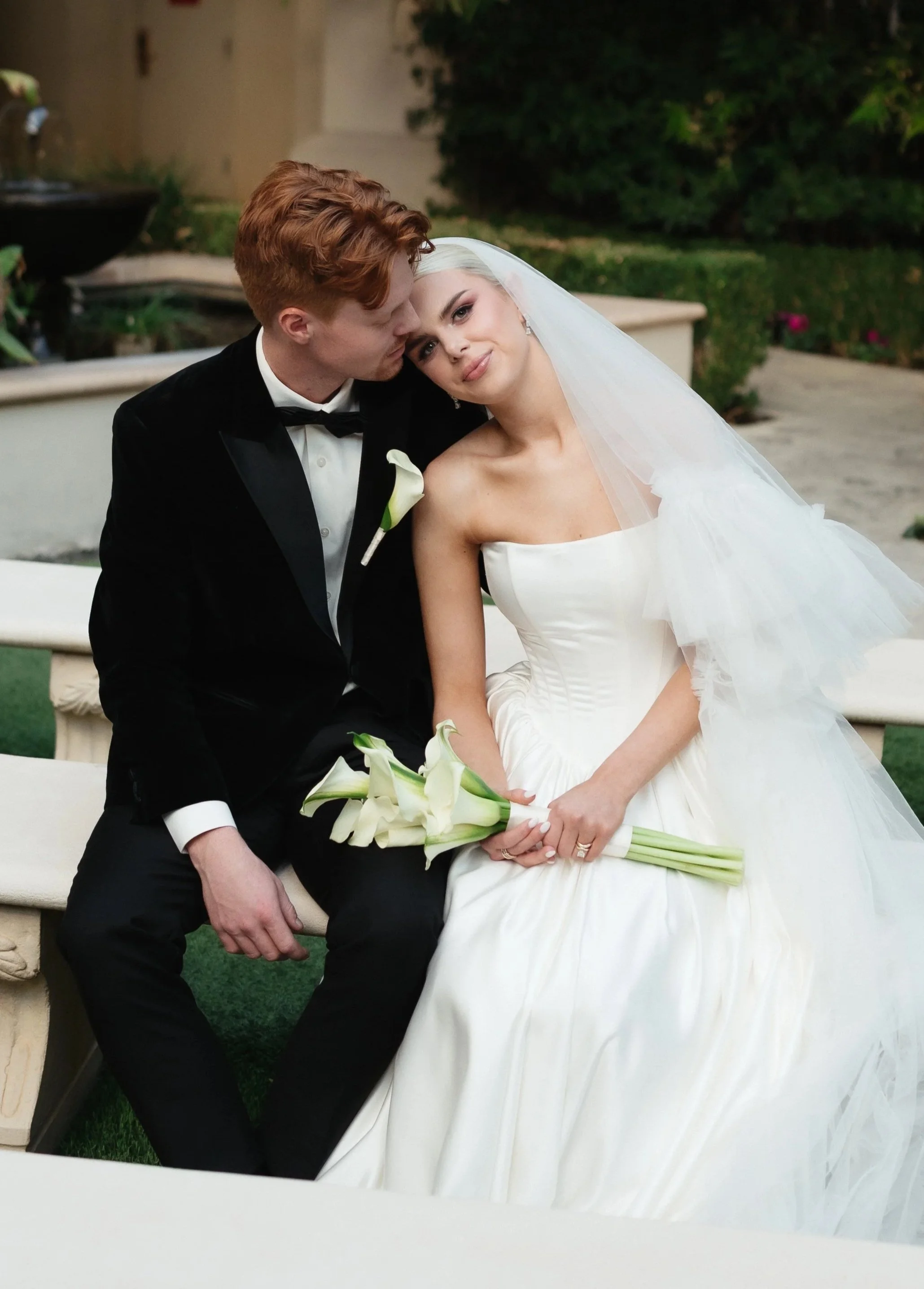 A bride and groom sitting closely on a bench during their wedding. The groom, with red hair and a black tuxedo, is leaning in to kiss the bride. The bride, with platinum blonde hair and a white strapless wedding gown, is holding a bouquet of calla li