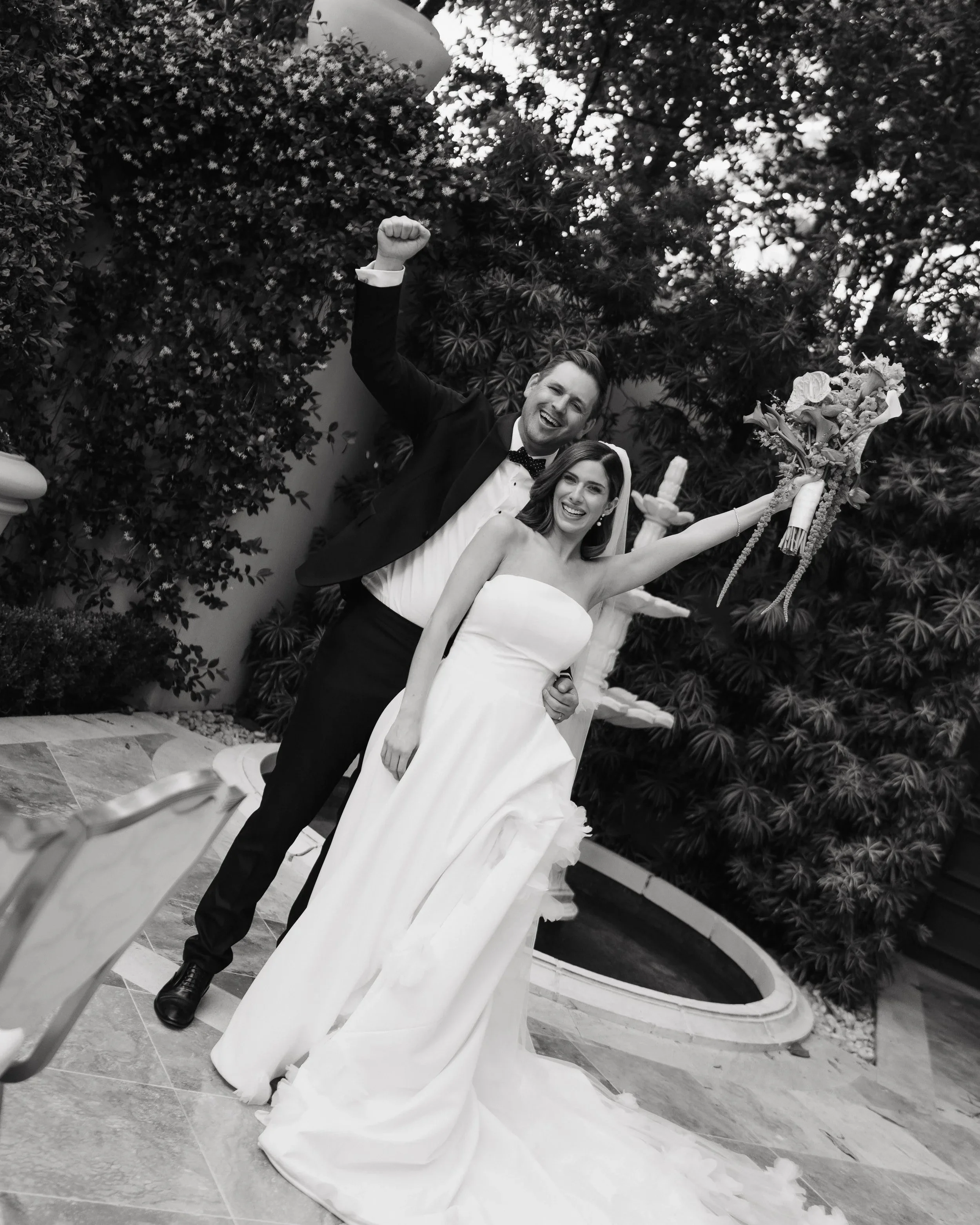 Black and white photo of a smiling bride and groom celebrating their wedding. The bride is in a white wedding dress holding a bouquet. The groom is in a tuxedo with a bowtie, raising his arm in celebration. They are outdoors with lush foliage in the 