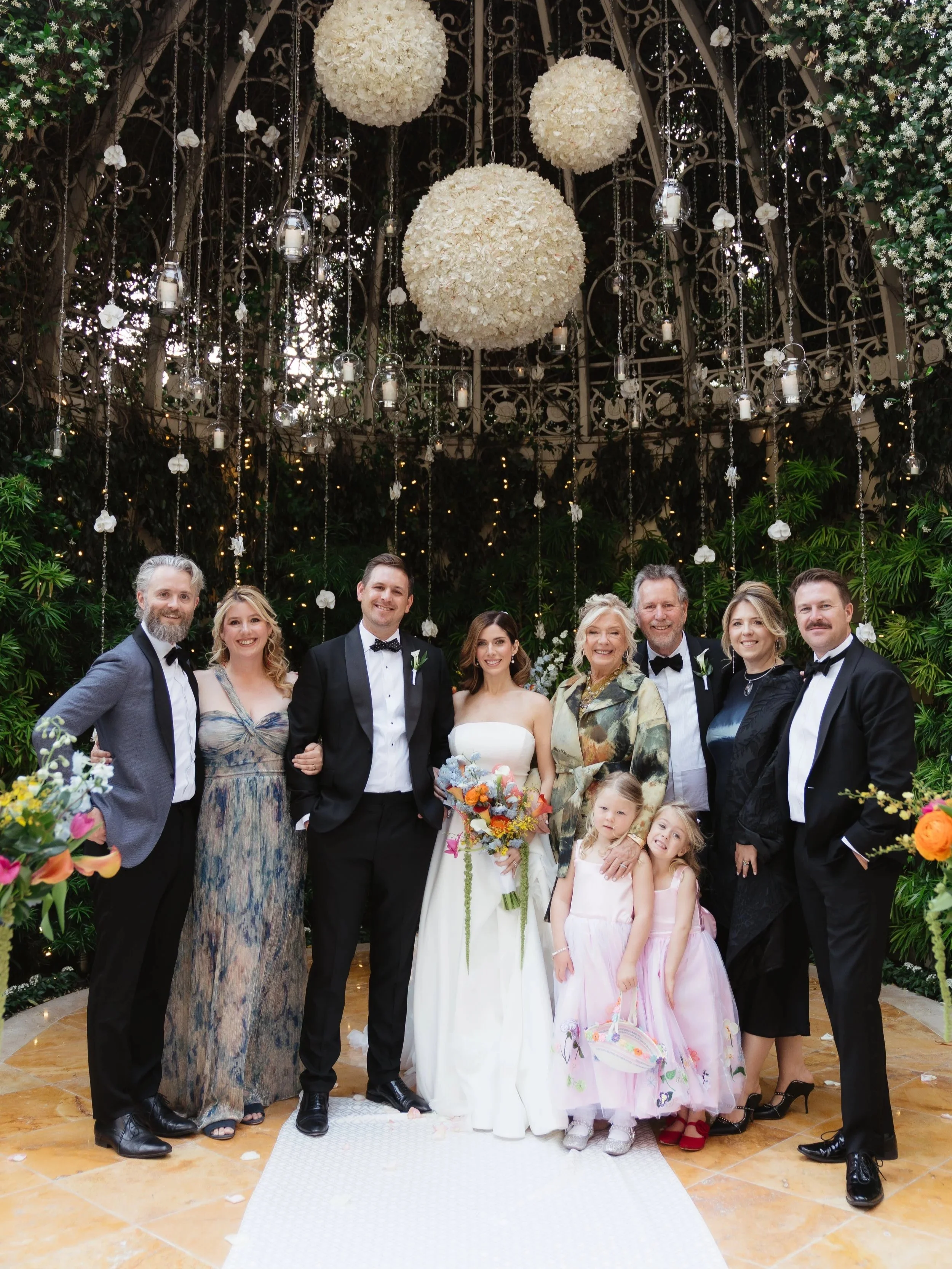 A group of people in formal attire at a wedding reception, standing under hanging white floral decorations and candles.