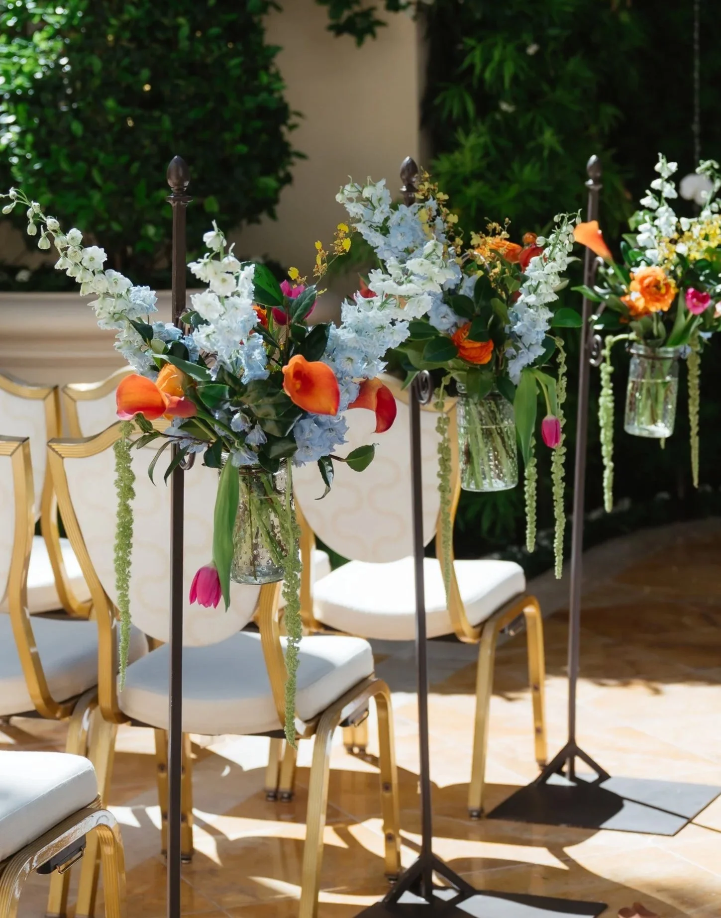 Flower arrangements with white, orange, pink, and yellow flowers in glass vases on black metal stands, set in a room with beige chairs and a beige couch, with green foliage in the background.