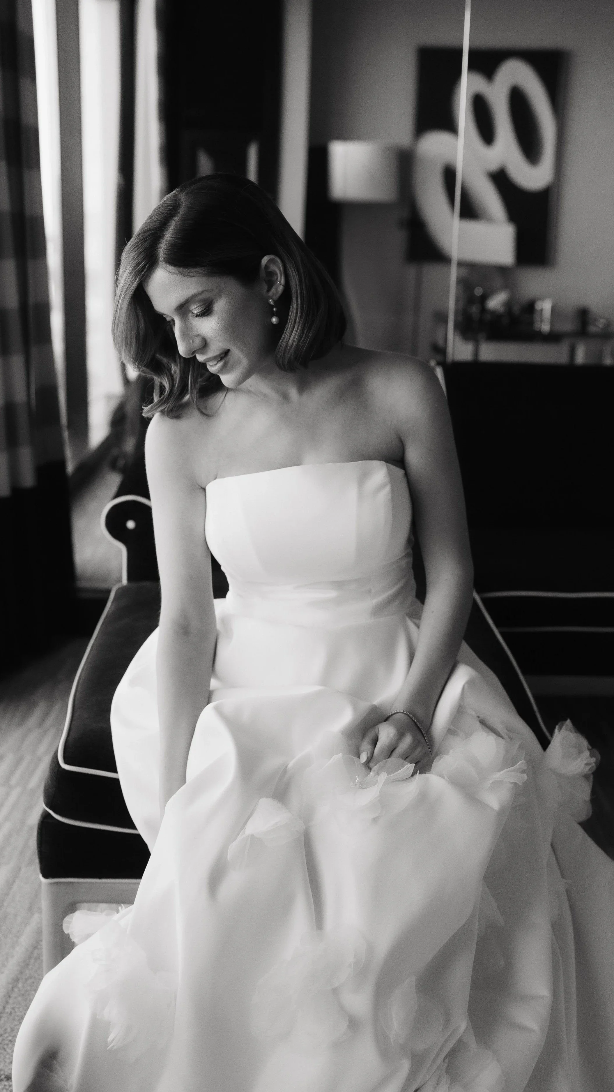 A woman wearing a strapless white wedding dress sitting on a chair, smiling and looking down, with a large abstract black and white painting in the background.