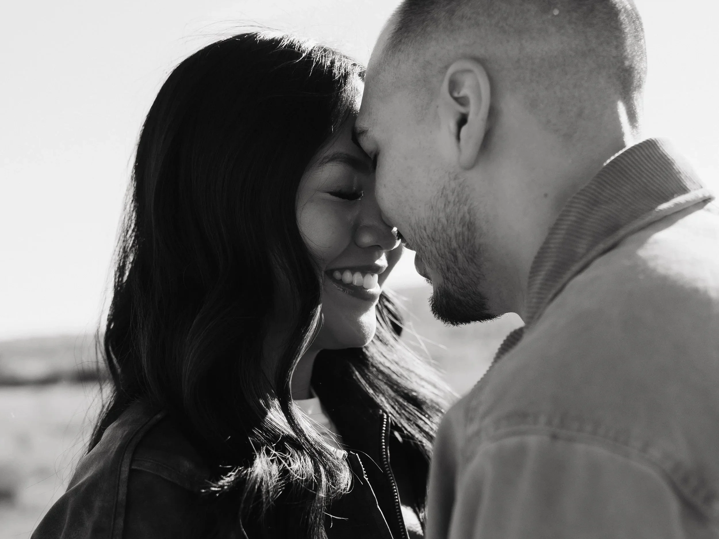 A black and white photo of a couple with foreheads touching, smiling, outdoors.