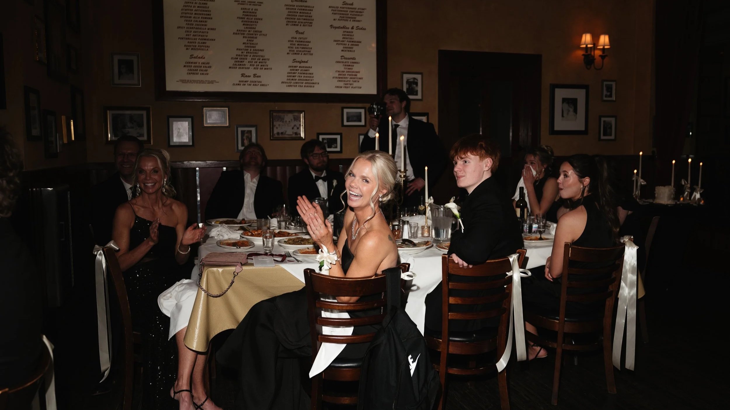 A group of people dressed in formal attire sitting around a dinner table at a restaurant, with a woman in the foreground smiling and clapping, others engaging in conversation, and a man standing in the background taking a photo. The restaurant has fr