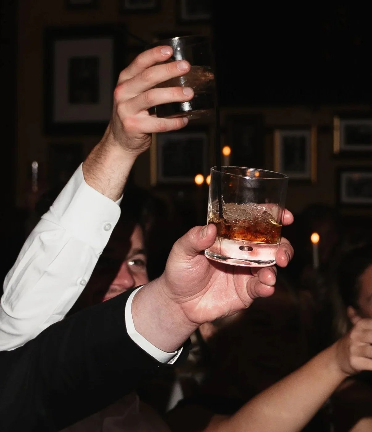 Two people toasting with glasses of whiskey and ice cubes in a dimly lit environment.