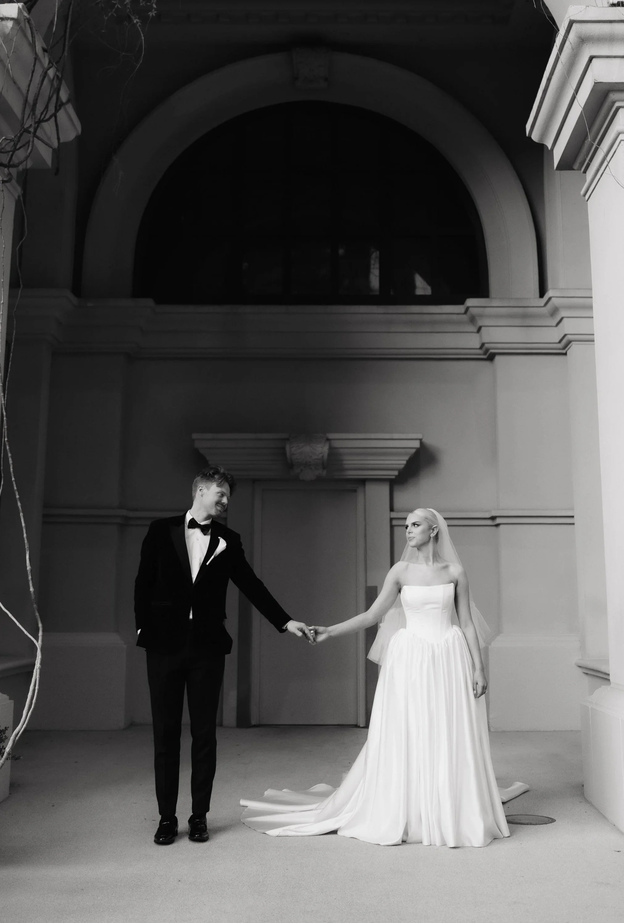 A bride and groom holding hands during a wedding ceremony, with the groom in a black tuxedo and the bride in a strapless white wedding gown, standing in an elegant indoor setting.