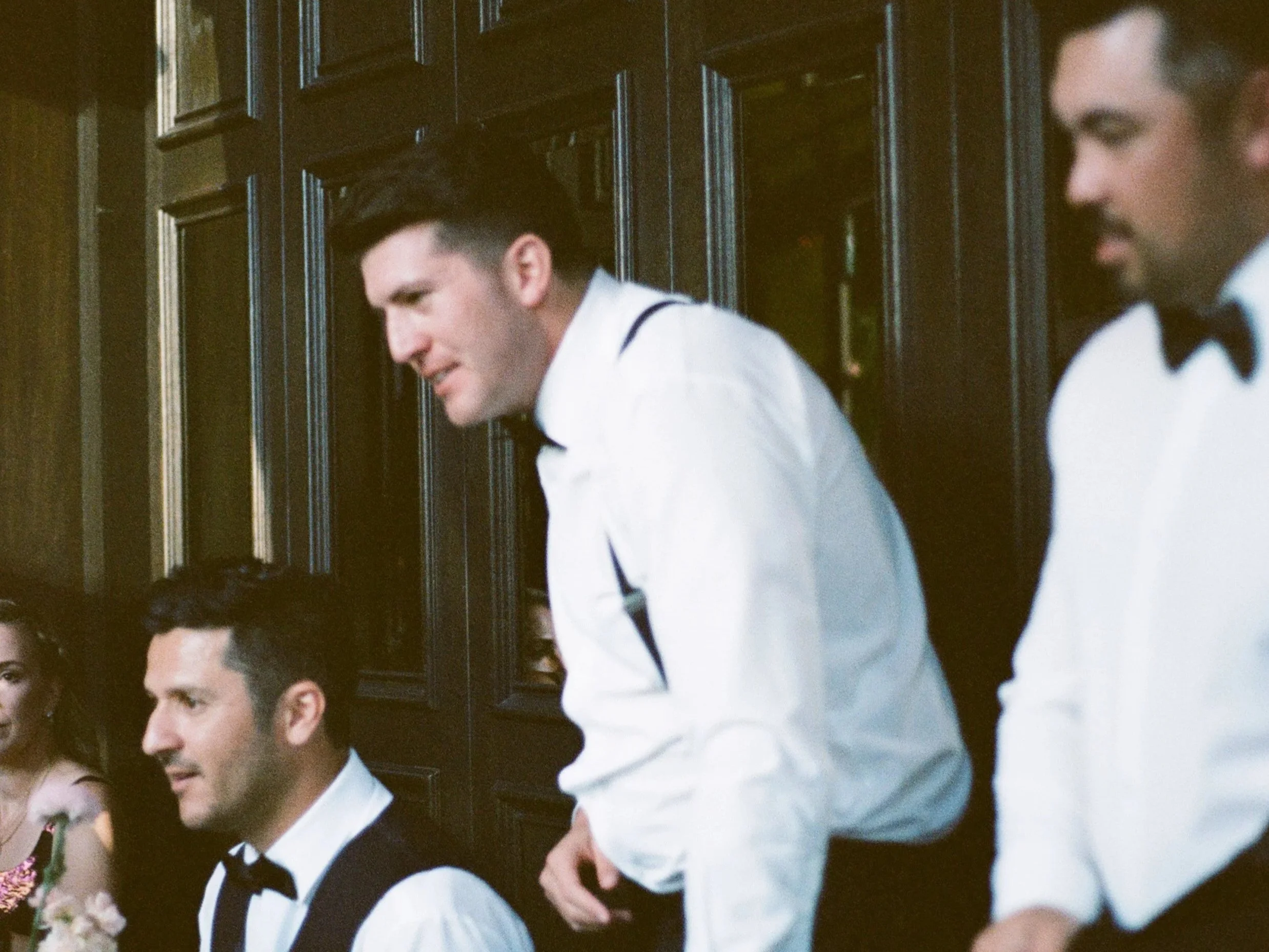 Group of people dressed in tuxedos and formal dresses celebrating at a wedding reception, with two men in tuxedos in focus, one smiling and the other looking serious, against a wooden-paneled wall.