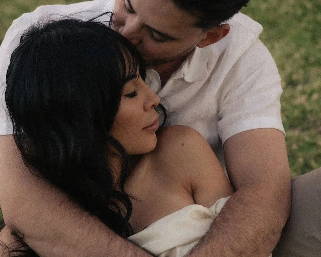 A romantic moment between a man and a woman, with the man embracing the woman from behind and kissing her forehead while she closes her eyes.